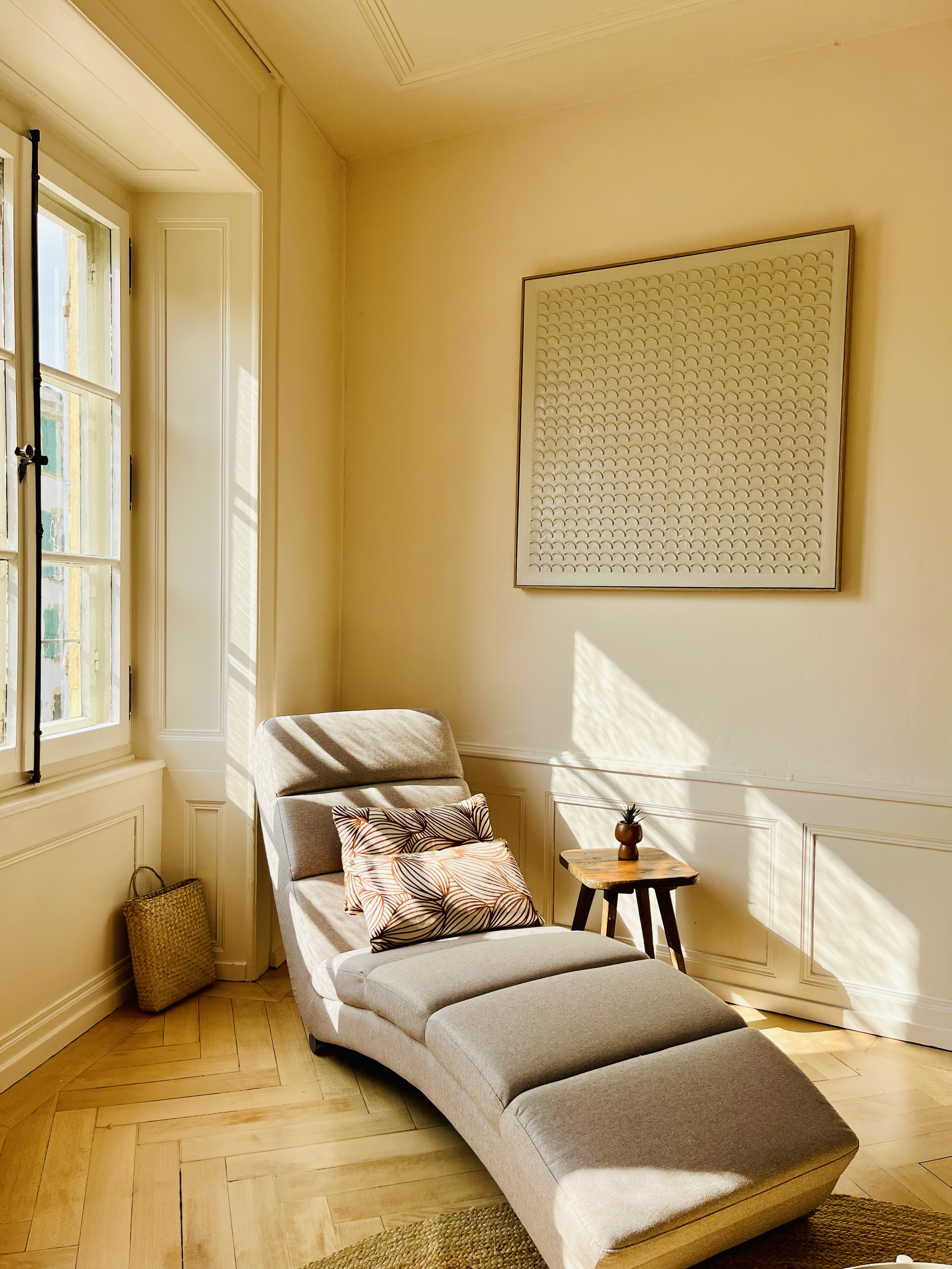 Une pièce lumineuse avec un fauteuil beige, un coussin à motif feuillage, une petite table en bois avec une plante en pot, une fenêtre laissant entrer la lumière, et un tableau abstrait au mur.