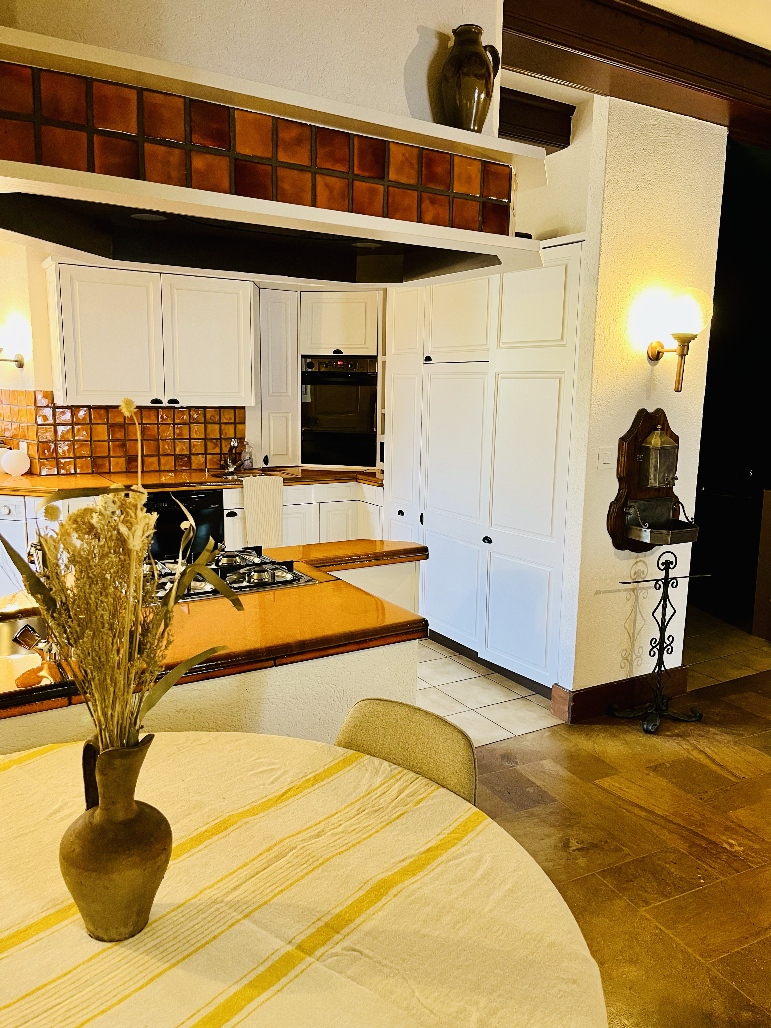 Cuisine rustique avec des armoires blanches, un plan de travail en bois, des carreaux de cuisine orange-brun, un four intégré, un dosseret en carreaux, un plafond en bois, une table avec une nappe beige et un vase avec des fleurs séchées.