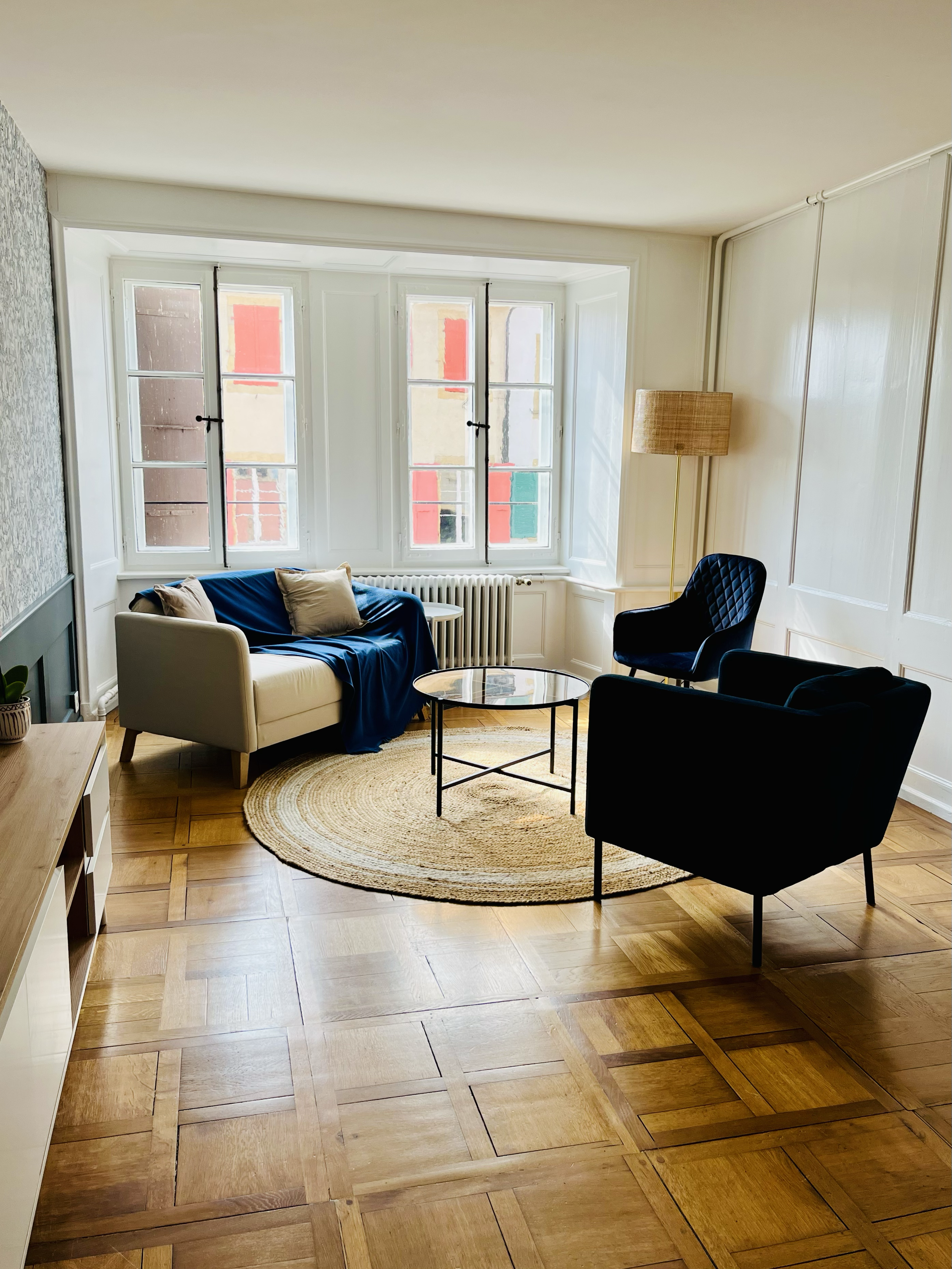Salon lumineux avec canapé beige, deux fauteuils noirs, un tapis rond en osier, une table basse ronde en verre, une lampe à piédestal et plusieurs fenêtres laissant entrer la lumière naturelle.