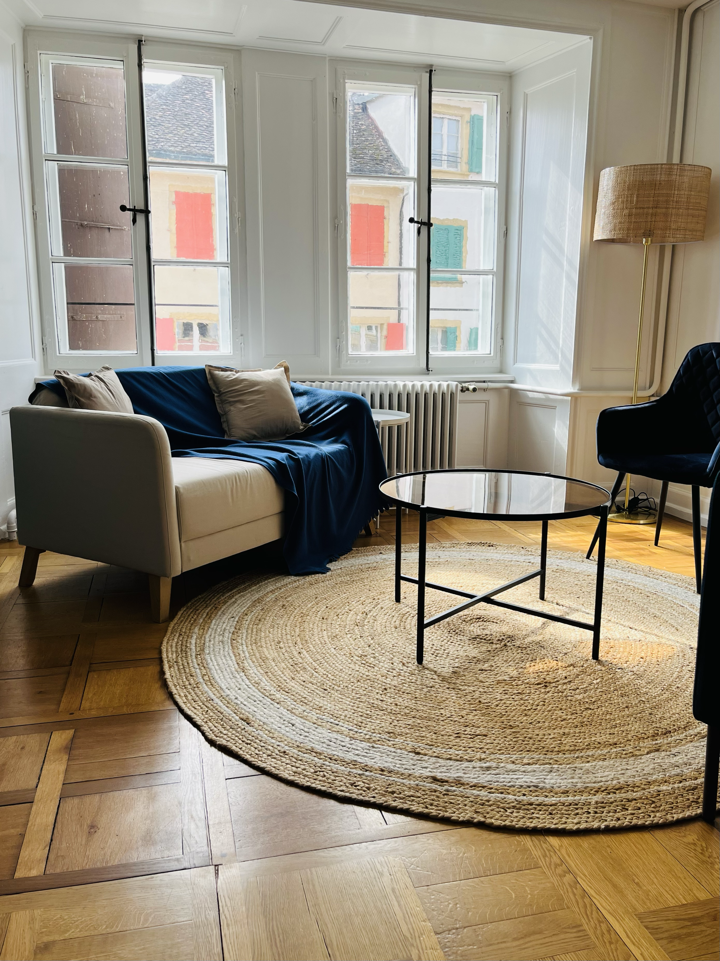Salon avec canapé beige, tapis rond en osier, table en verre, chaise bleue, lampadaire en rotin, fenêtres avec vue sur des bâtiments colorés.