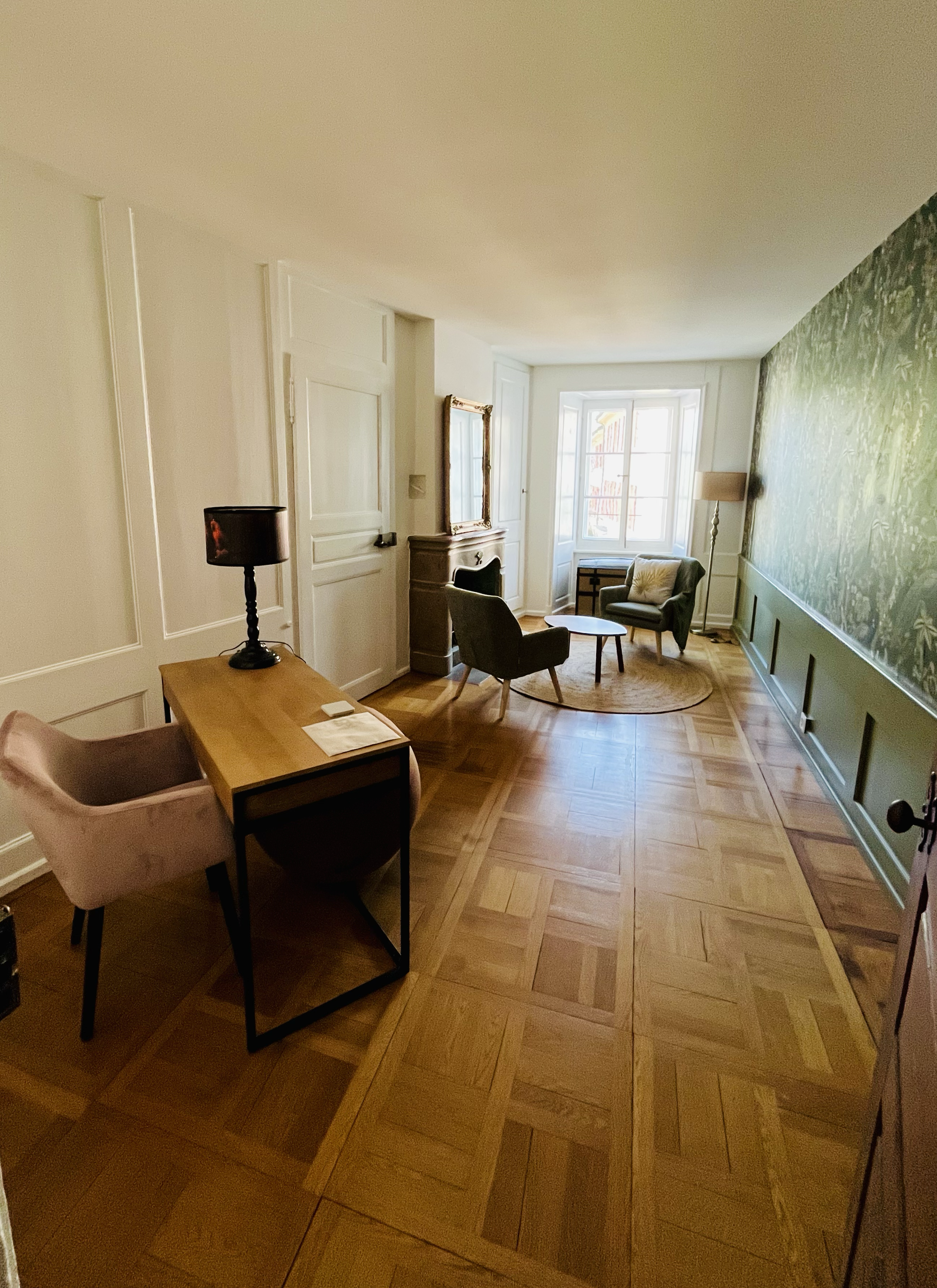 Salon avec parquet en bois clair, fenêtre laissant entrer la lumière naturelle, fauteuils verts, table basse, lampe de sol, cheminée ancienne, mur vert avec motif floral, bureau en bois avec lampe noire.