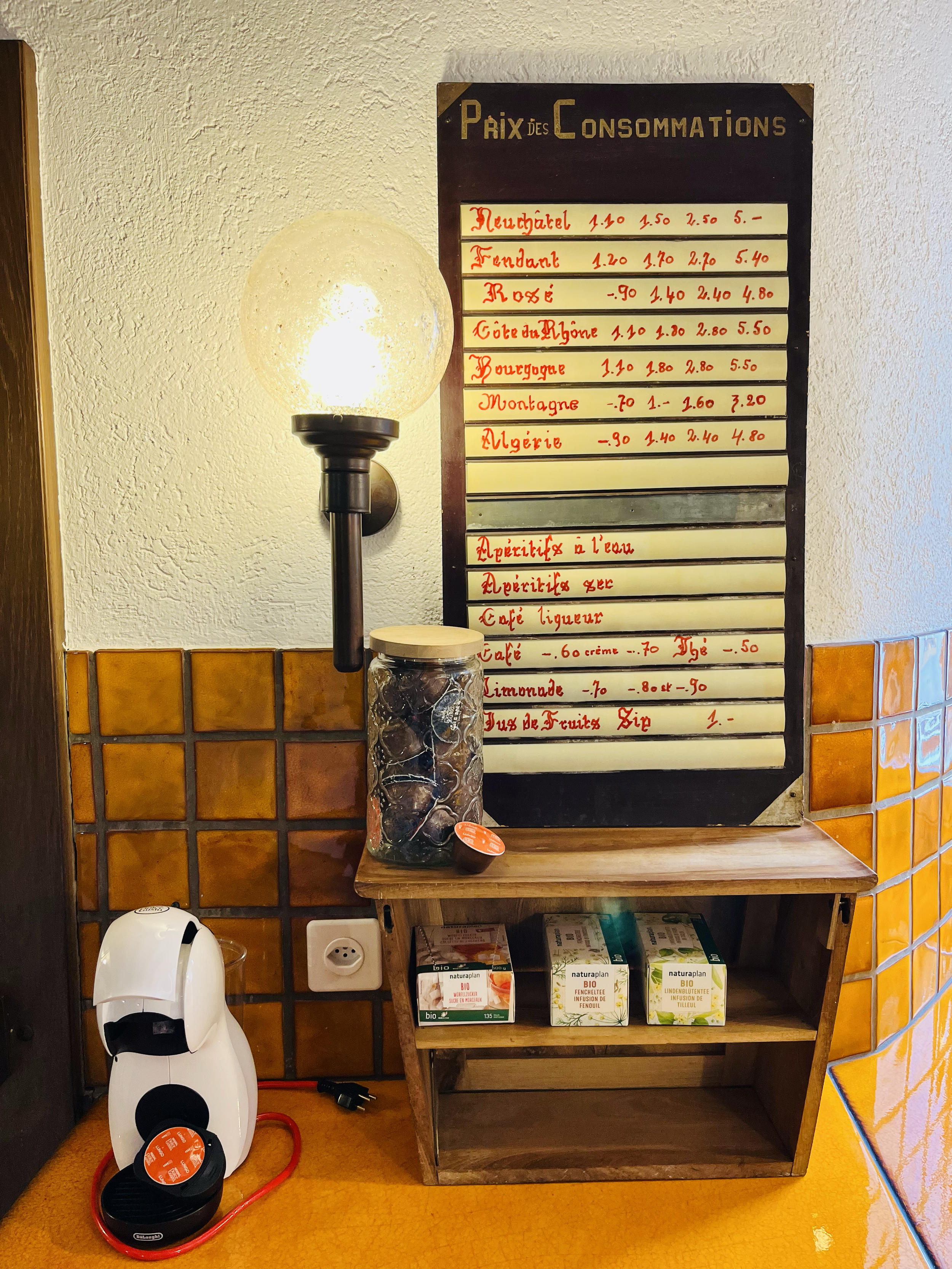 Tableau indiquant les prix des boissons et spécialités, avec une lampe à gauche, une jarre de ce qui semble être des olives ou des fruits secs, et une petite étagère contenant des boîtes de thé biologiques.»
