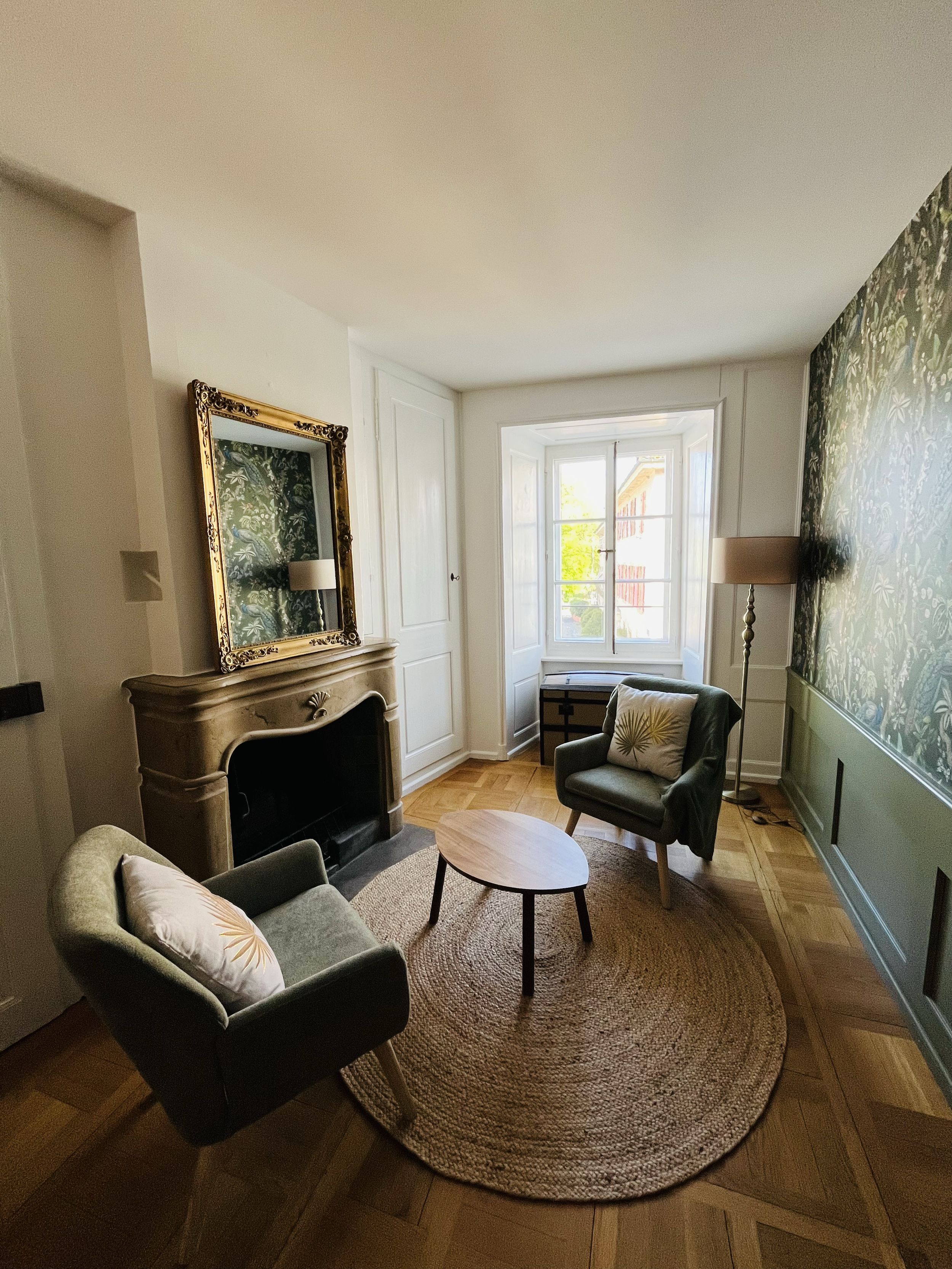 Salon avec deux fauteuils, tapis rond en jute, cheminée en pierre, miroir orné, grande fenêtre laissant entrer la lumière, tapisserie florale sur un mur, lampe sur pied.