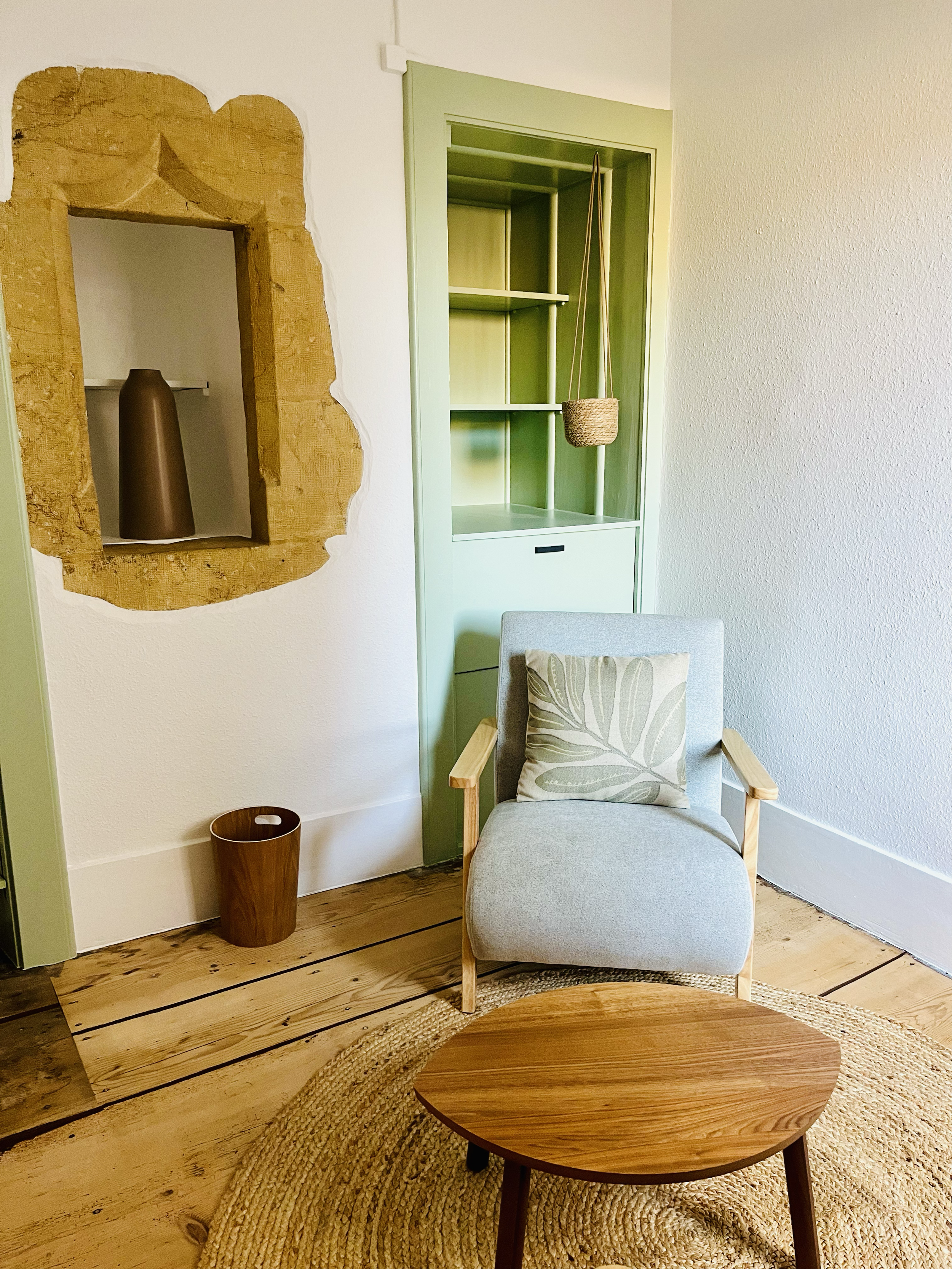 Salon avec une chaise en tissu gris, un coussin à motif de feuille, une table en bois ovale, un mur avec une niche en pierre avec une vase marron, une étagère en vert pâle avec une panière suspendue, sur un sol en bois et un tapis rond en fibres natu