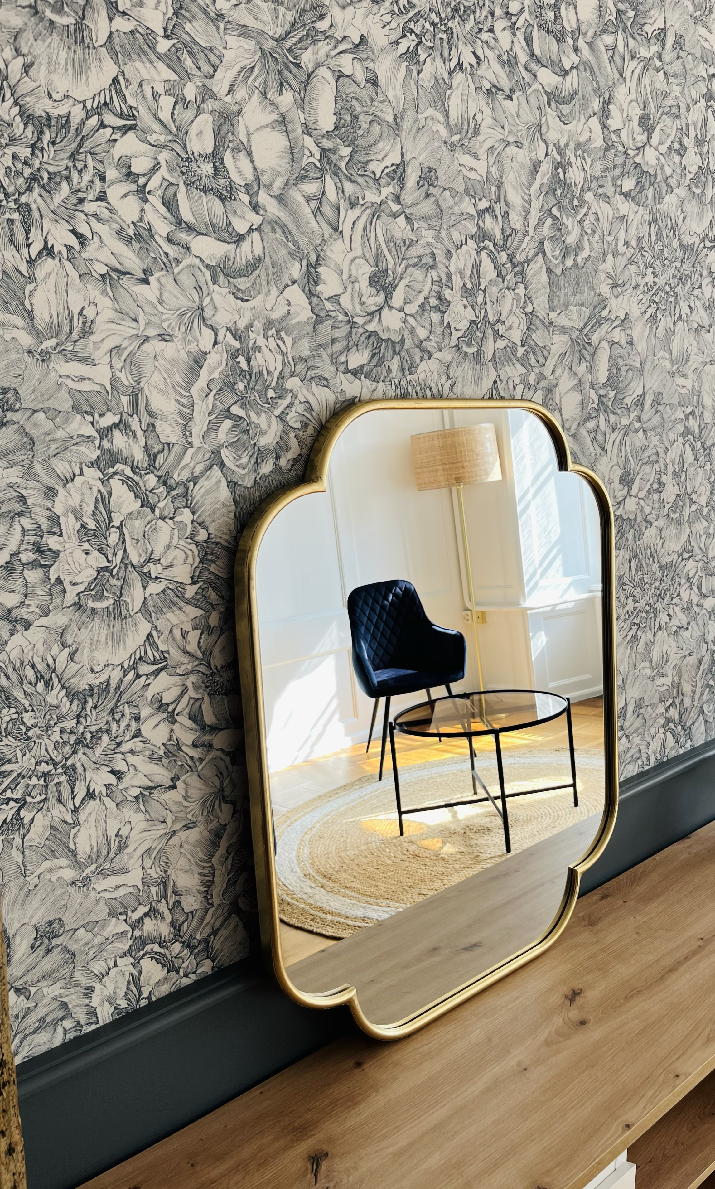 Un secrétaire en bois avec un miroir mural orné d'un cadre doré représentant un fauteuil noir, un tapis rond beige, une lampe sur pied, dans une pièce lumineuse avec murs en bois blanc et un mur tapissé de papier peint floral en noir et blanc.