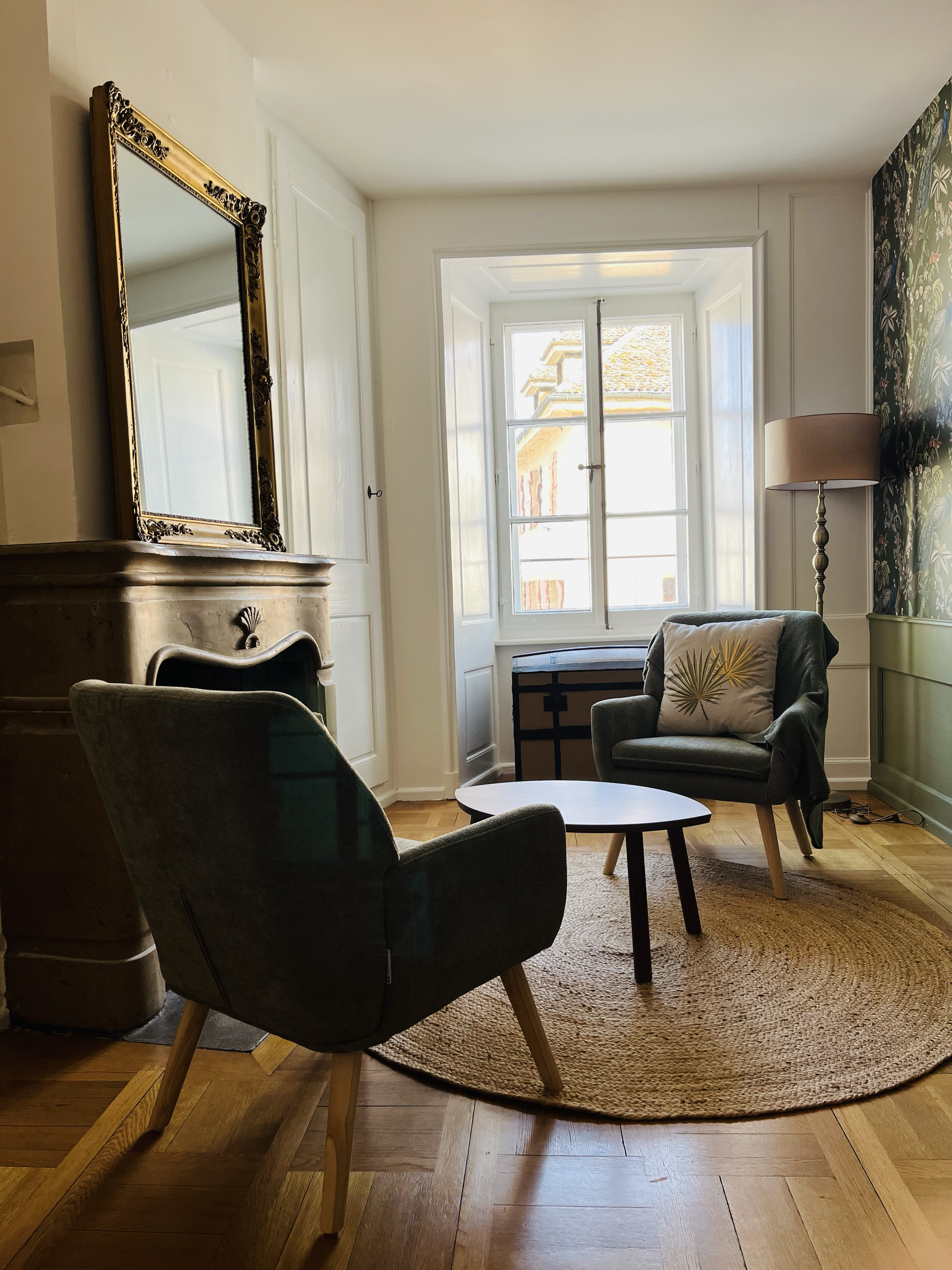 Salon avec deux fauteuils en tissu vert, un petit tapis rond en jute, un coffre à lierre, une fenêtre, un lampadaire sur pied et un miroir ancien au-dessus d'une cheminée en marbre.