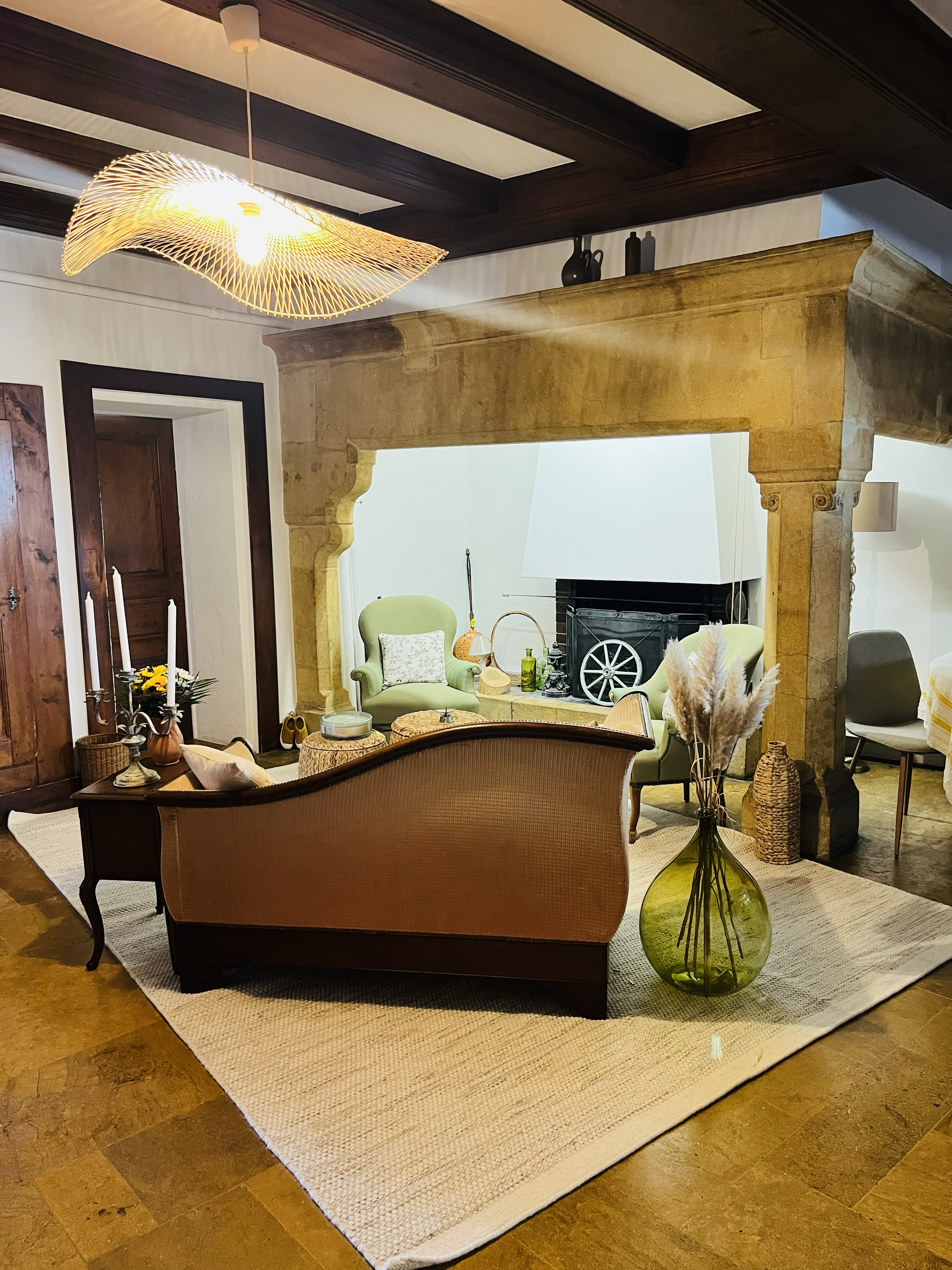 Intérieur d’un salon avec un canapé, deux fauteuils verts, un tapis beige, une cheminée, une grande jarre verte avec des fleurs séchées, et des décorations en bois et céramique.
