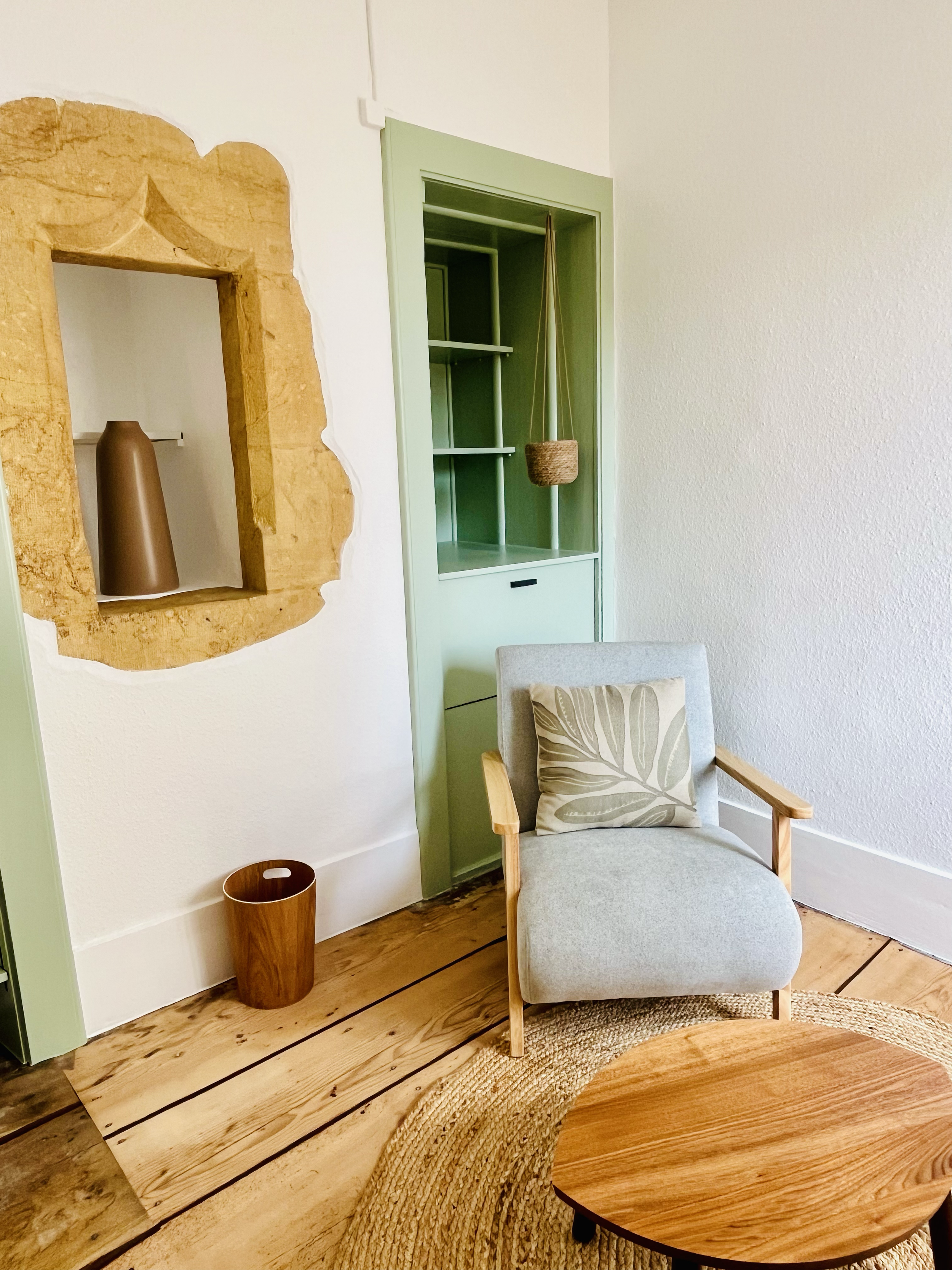 Coin salon avec un fauteuil gris clair, un coussin à motif de feuilles, une étagère verte encadrée, un plateau de table ronde en bois, un tapis en fibre naturelle, une niche murale en pierre contenant un vase marron et une petite boîte en osier suspe