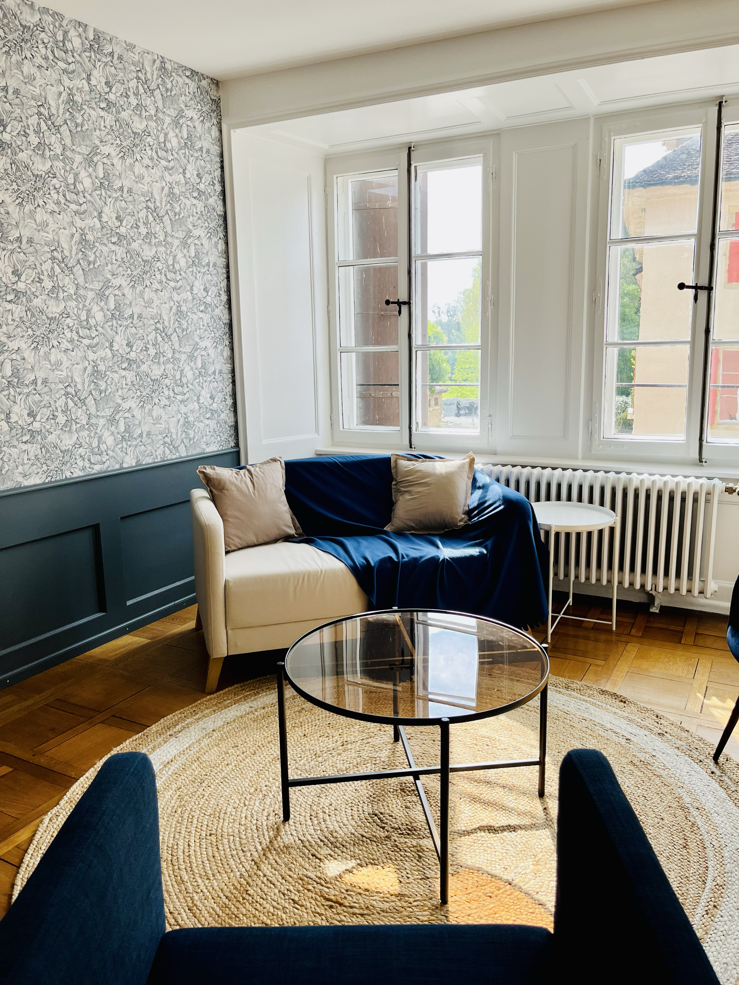 Salon lumineux avec canapé blanc, coussins beige, plaid bleu, table en verre, fauteuil bleu foncé, mur avec papier peint floral gris et blanc, grand fenêtre avec vue sur bâtiments et arbres, parquet en bois, tapis rond en jute.