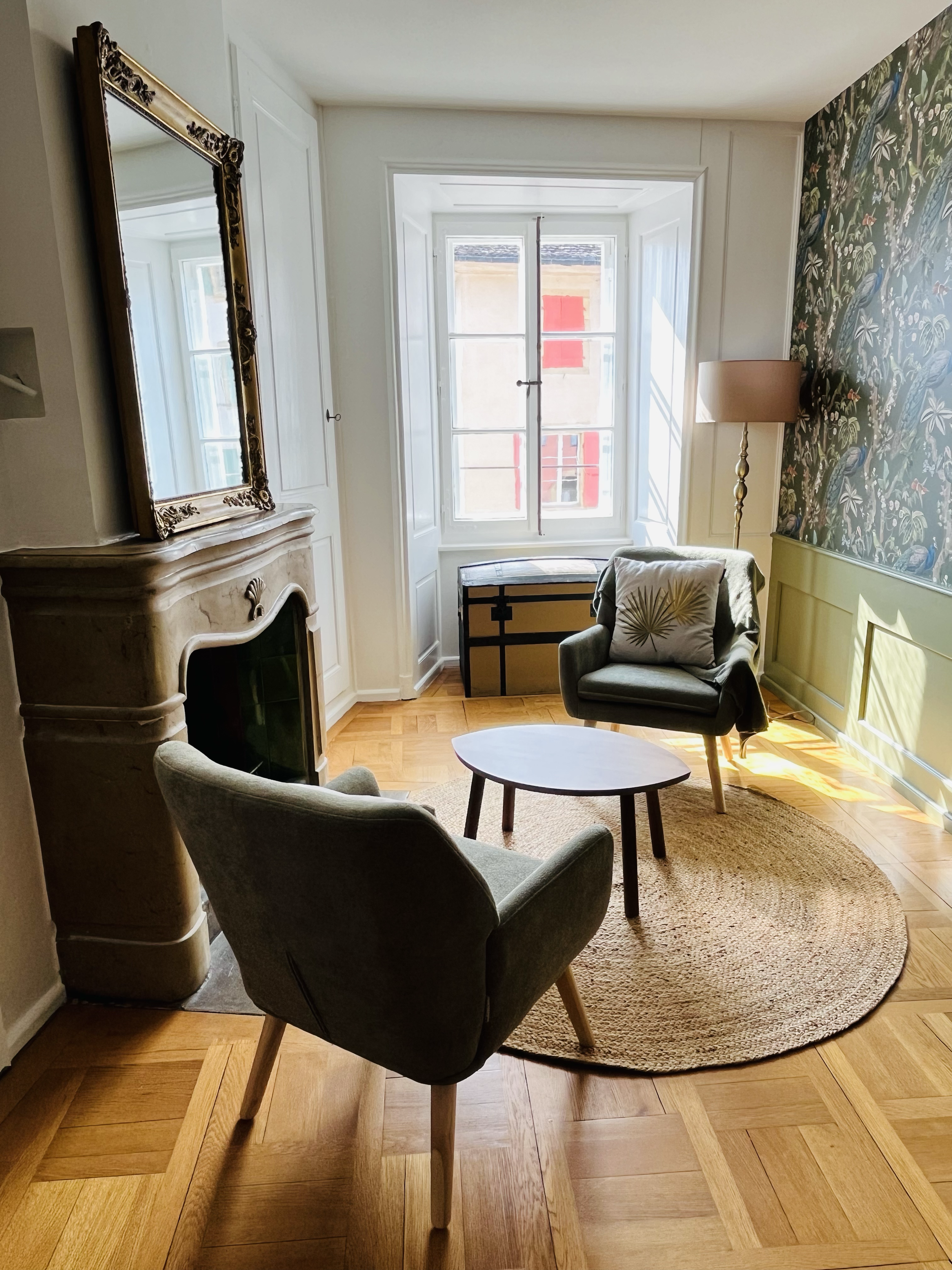 Salon avec cheminée en pierre, deux fauteuils, une table ronde, une lampe sur pied, un coin de tapis rond, un coffre sous une fenêtre avec volets rouges, mur tapissé avec motifs de perroquets, et un grand miroir
