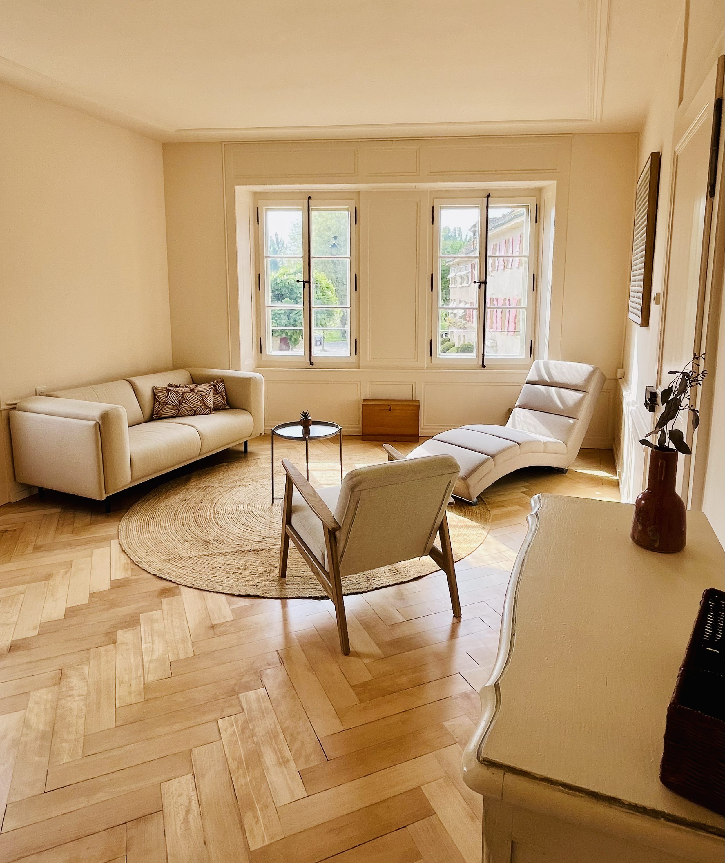 Salon lumineux avec canapés blancs, fauteuils, une table basse ronde, et des fenêtres laissant entrer la lumière naturelle. Plancher en bois et décoration minimaliste.