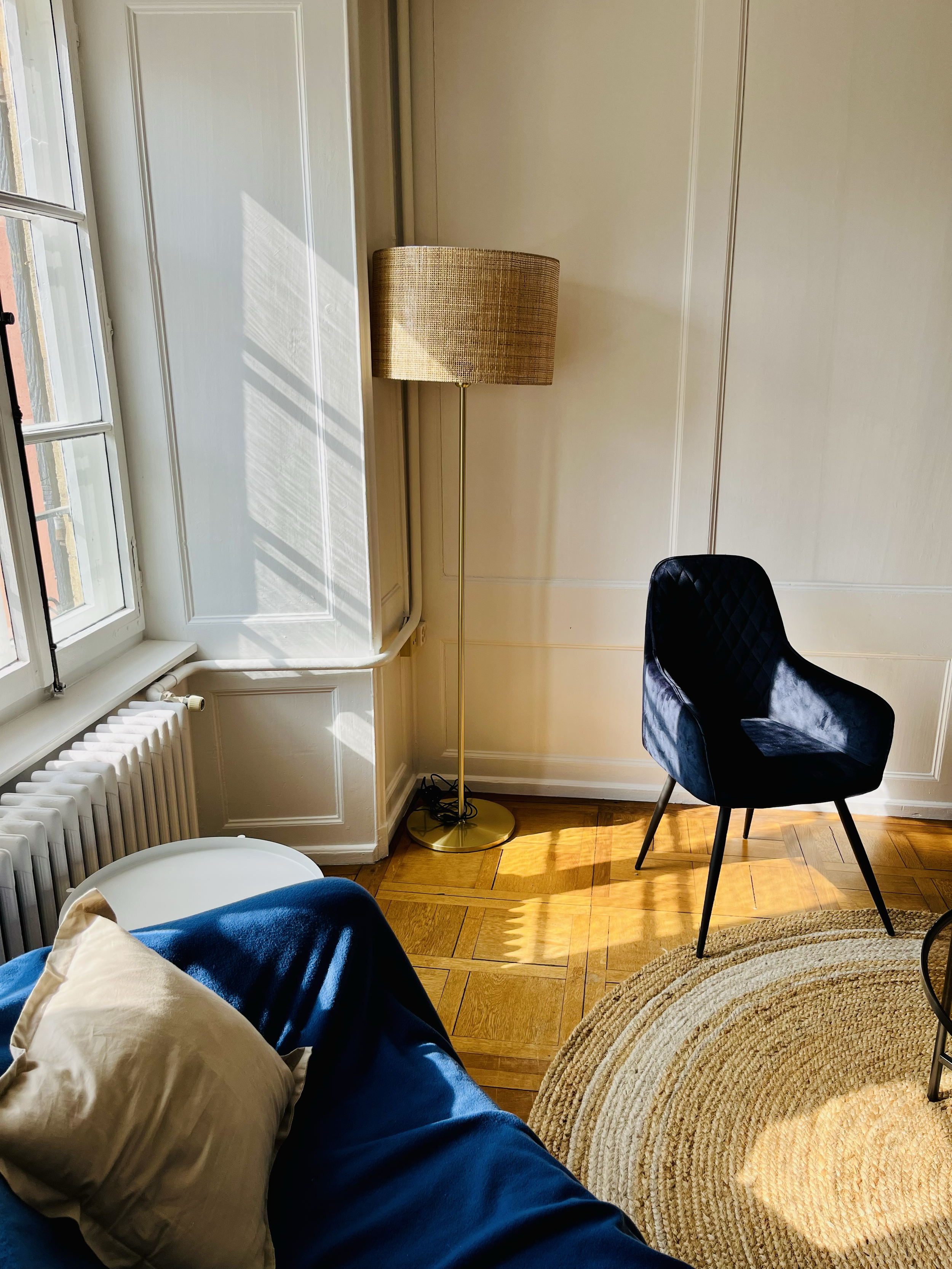 Coin salon avec un canapé bleu, un coussin beige, un fauteuil noir, une lampe sur pied, un tapis rond en jute, un radiateur et une fenêtre laissant entrer la lumière