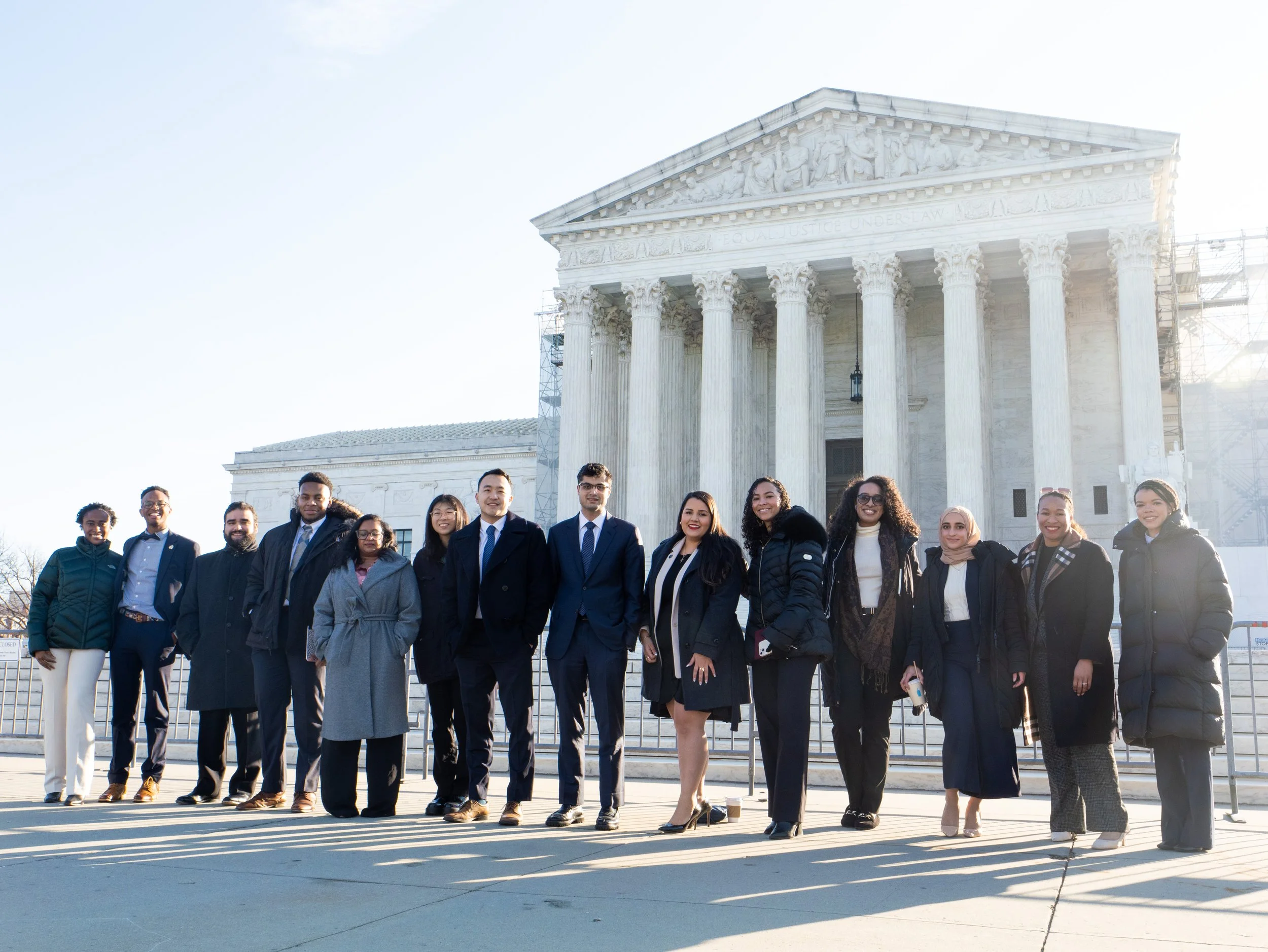 A diverse group of people dressed in professional attire stand together in front of the United States Supreme Court building, smiling in the sunlight with the court’s white columns rising behind them.