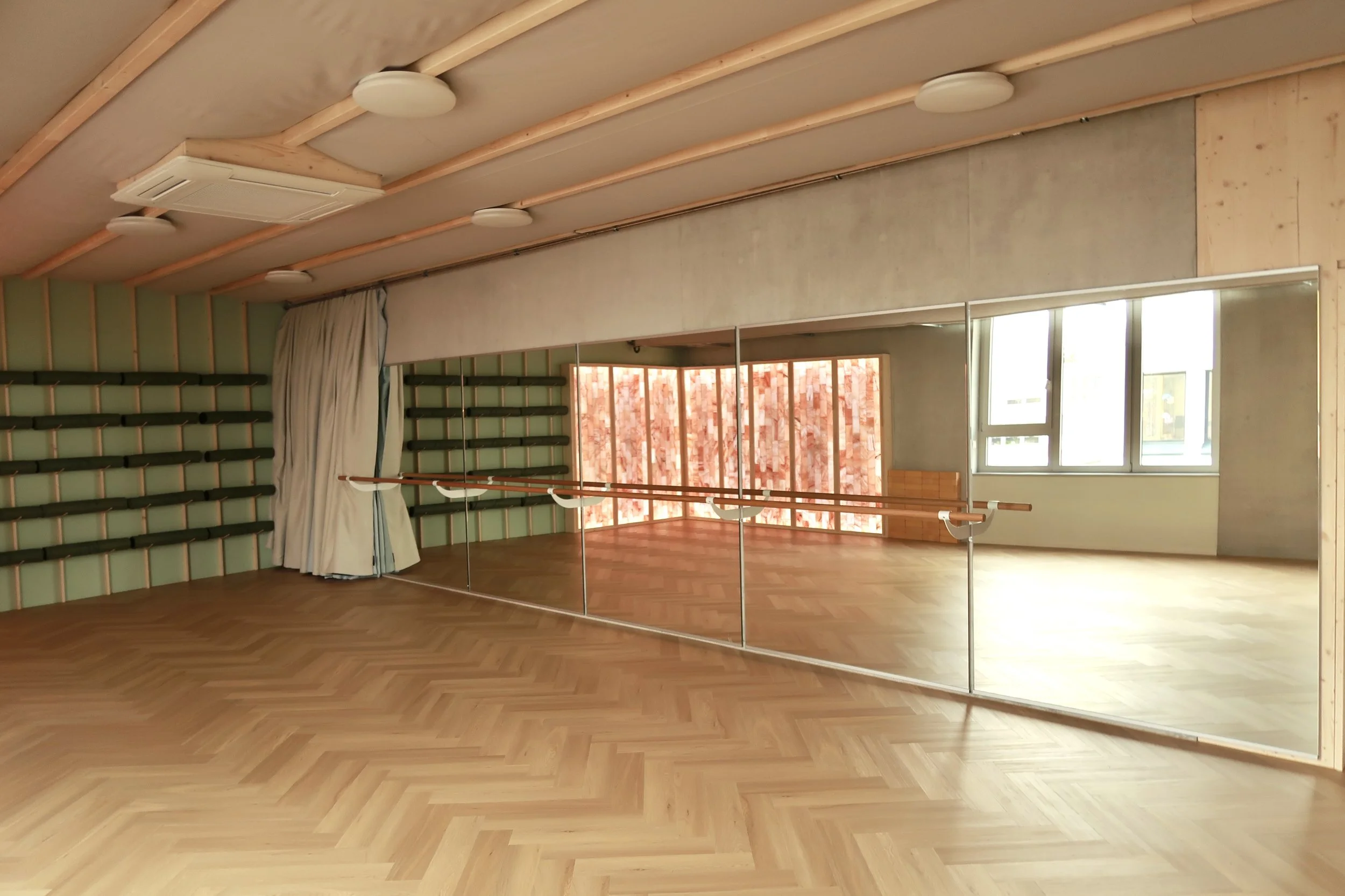 Empty dance and yoga studio with wooden floors, a large mirror, ballet barre, yoga mats, windows, and curtains.