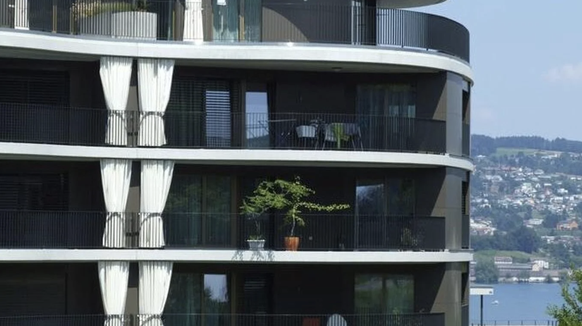 Mehrstöckiges modernes Wohngebäude mit mehreren Balkonen, teilweise mit weißen Vorhängen und Pflanzen, mit Wasser und Stadt im Hintergrund.