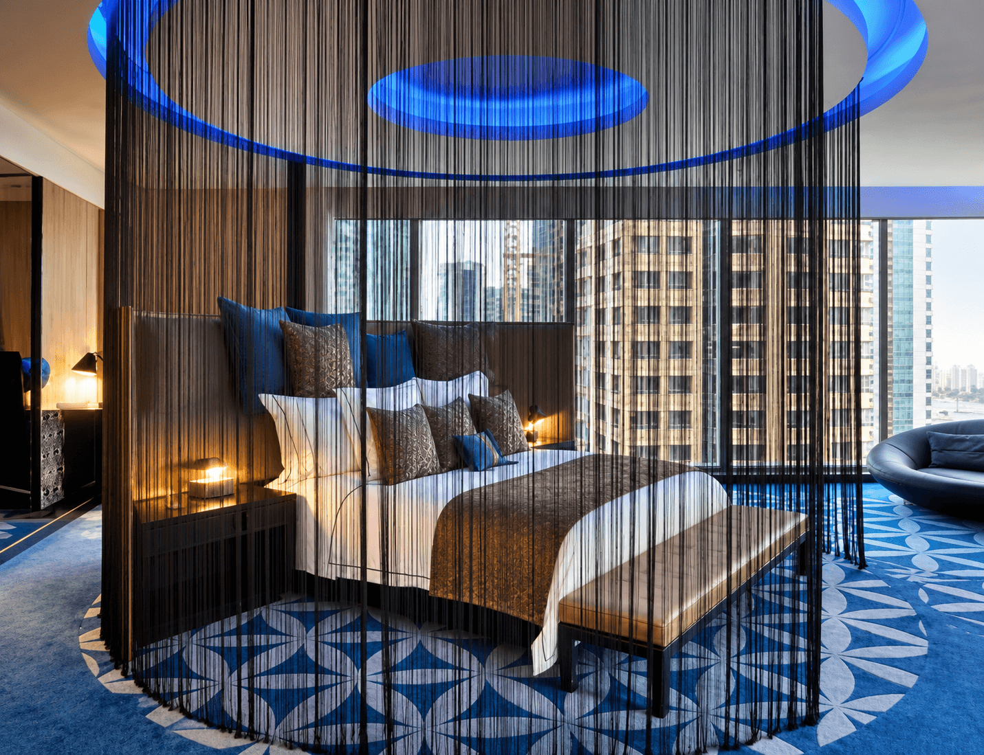 Ein modernes Hotelzimmer mit Bett, Zierkissen, Vorhängen und Stadtblick durch die Fenster.