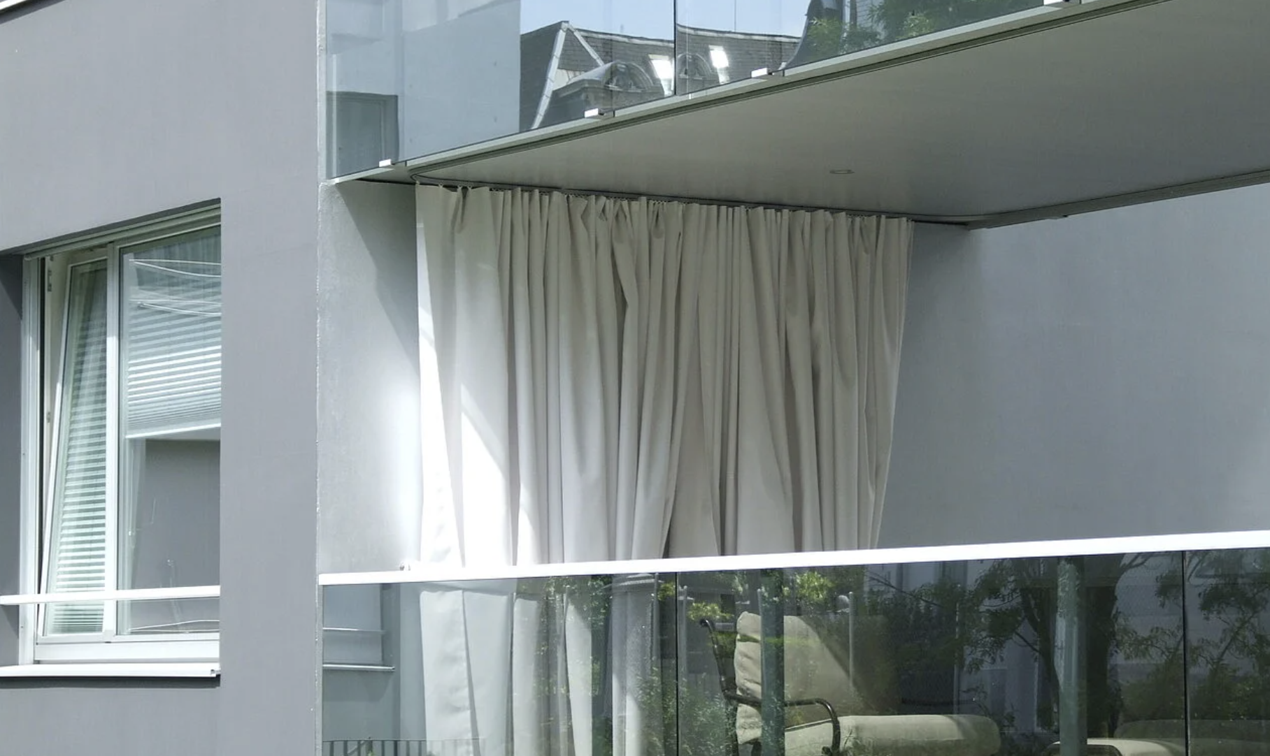 Moderne Gebäude mit großen Fenstern, weißen Vorhängen und einer Terrasse mit Glasbalustrade.