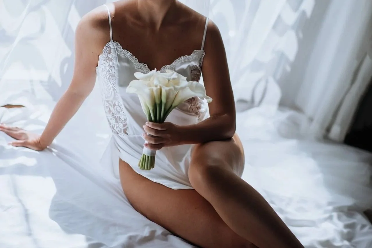 Bride in white bridal lingerie holding a bouquet while sitting on a bed