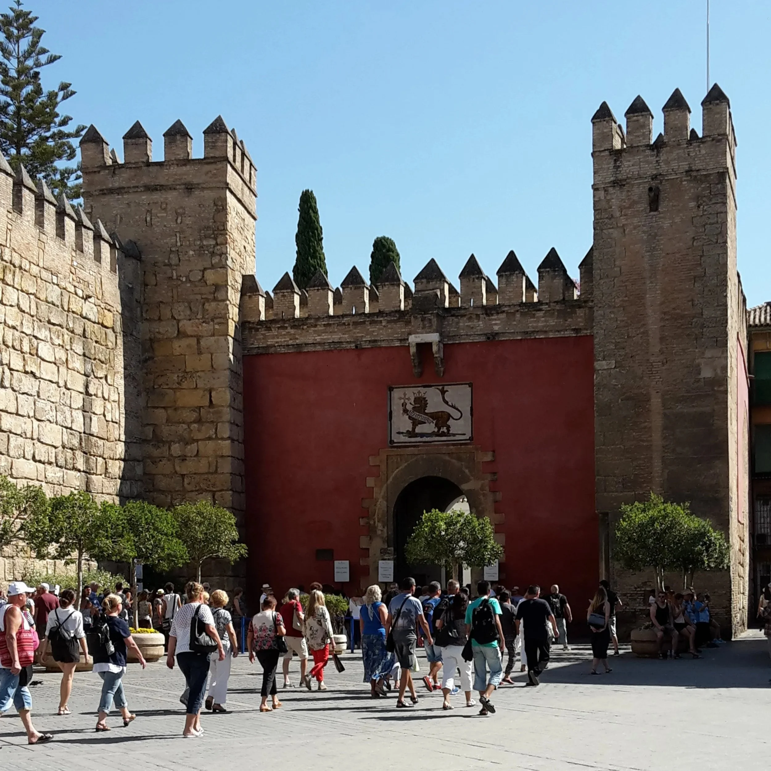 Porte d'entrée d'une vieille forteresse avec une muraille en pierre et des tours, représentant un symbole médiéval, avec des gens en visite et des arbres dans la cour.