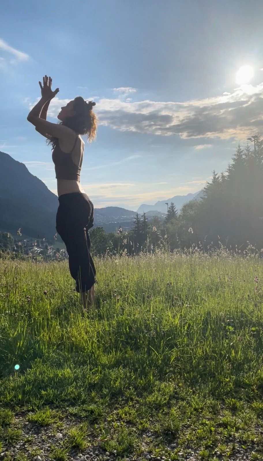 A person practicing yoga outdoors on a grassy field during sunset, with mountains in the background and the sun shining in the sky.