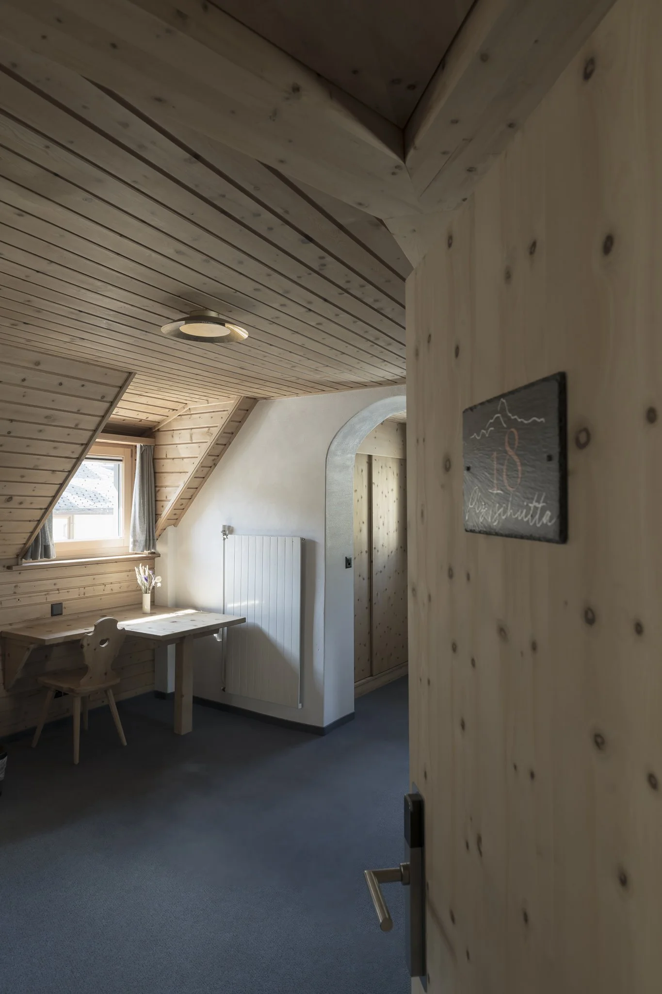 Interior of a cozy room with wooden ceiling and walls, small window with curtains, wooden desk with a chair, and a white radiator.