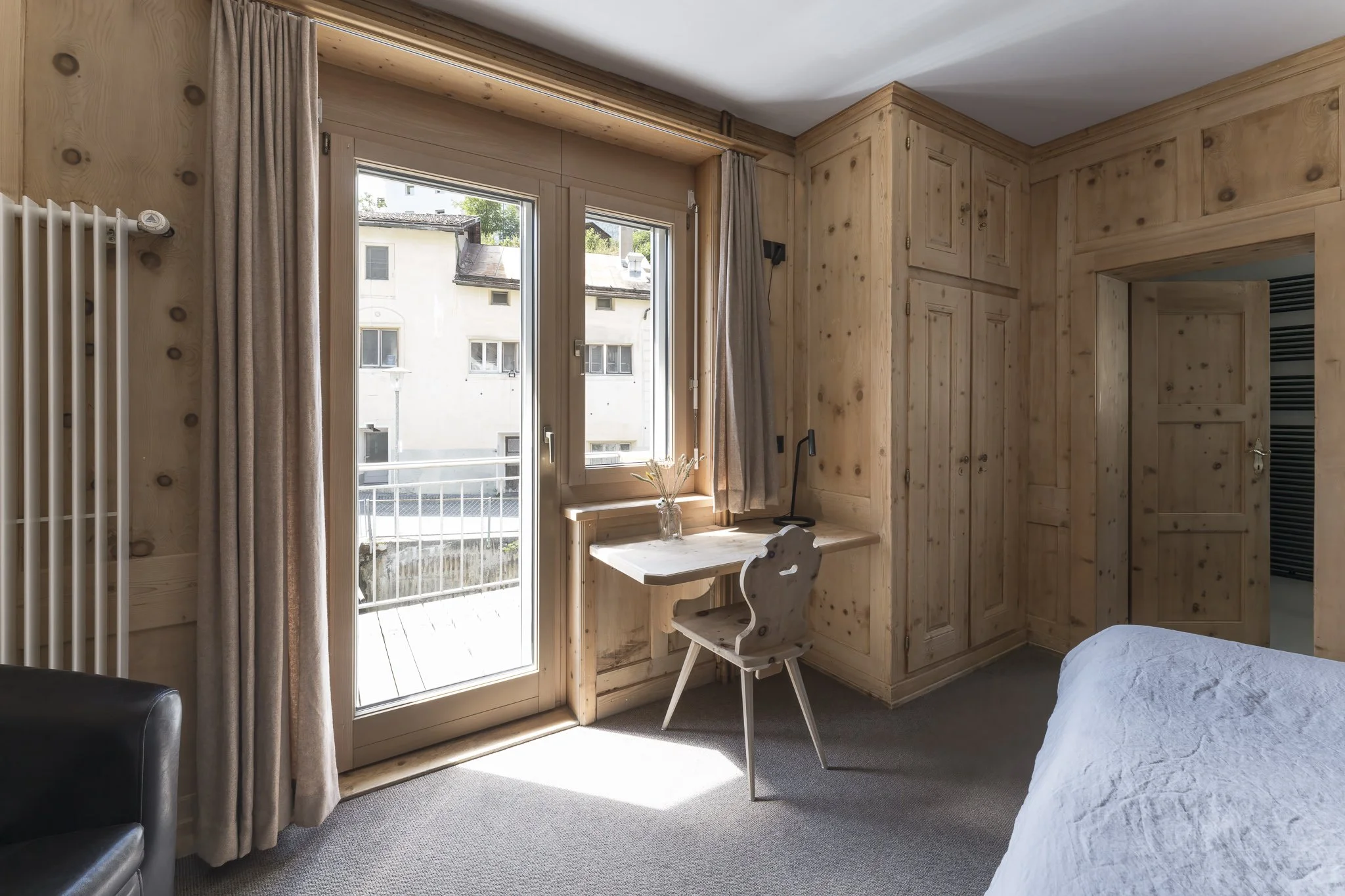 A cozy bedroom corner featuring wooden wall paneling, a window with beige curtains, a small desk with a chair, and a view of neighboring buildings outside.
