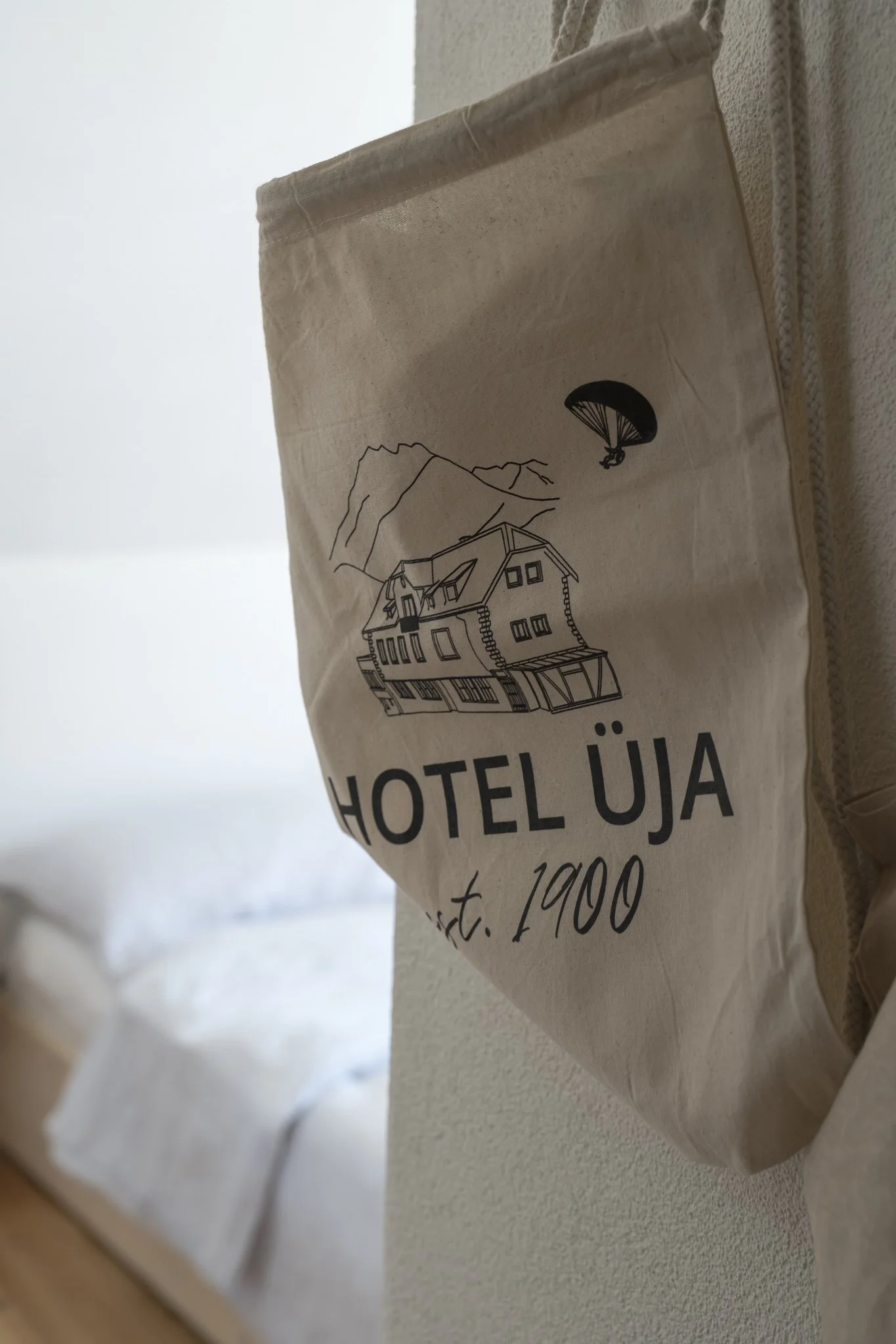 A beige fabric bag hanging on a wall, with a black line drawing of a building, mountain, and parachute, and the words 'HOTEL ÜJA est. 1900' printed on it.