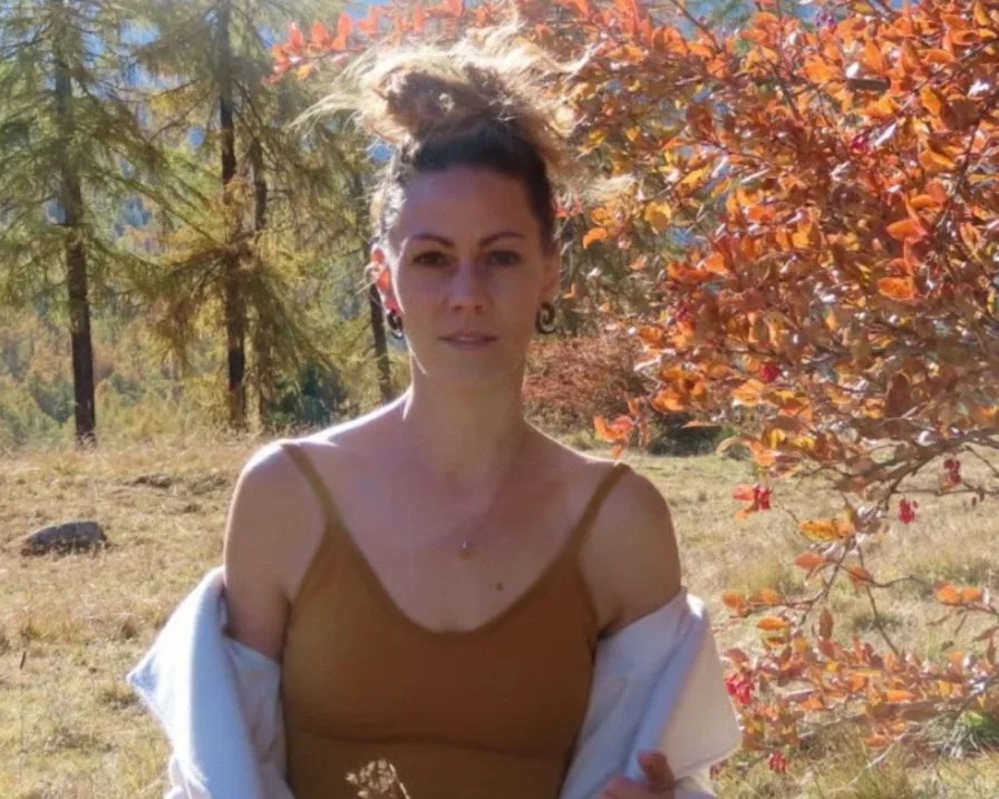 A woman with braided hair and earrings standing outdoors with autumn-colored leaves and trees in the background.