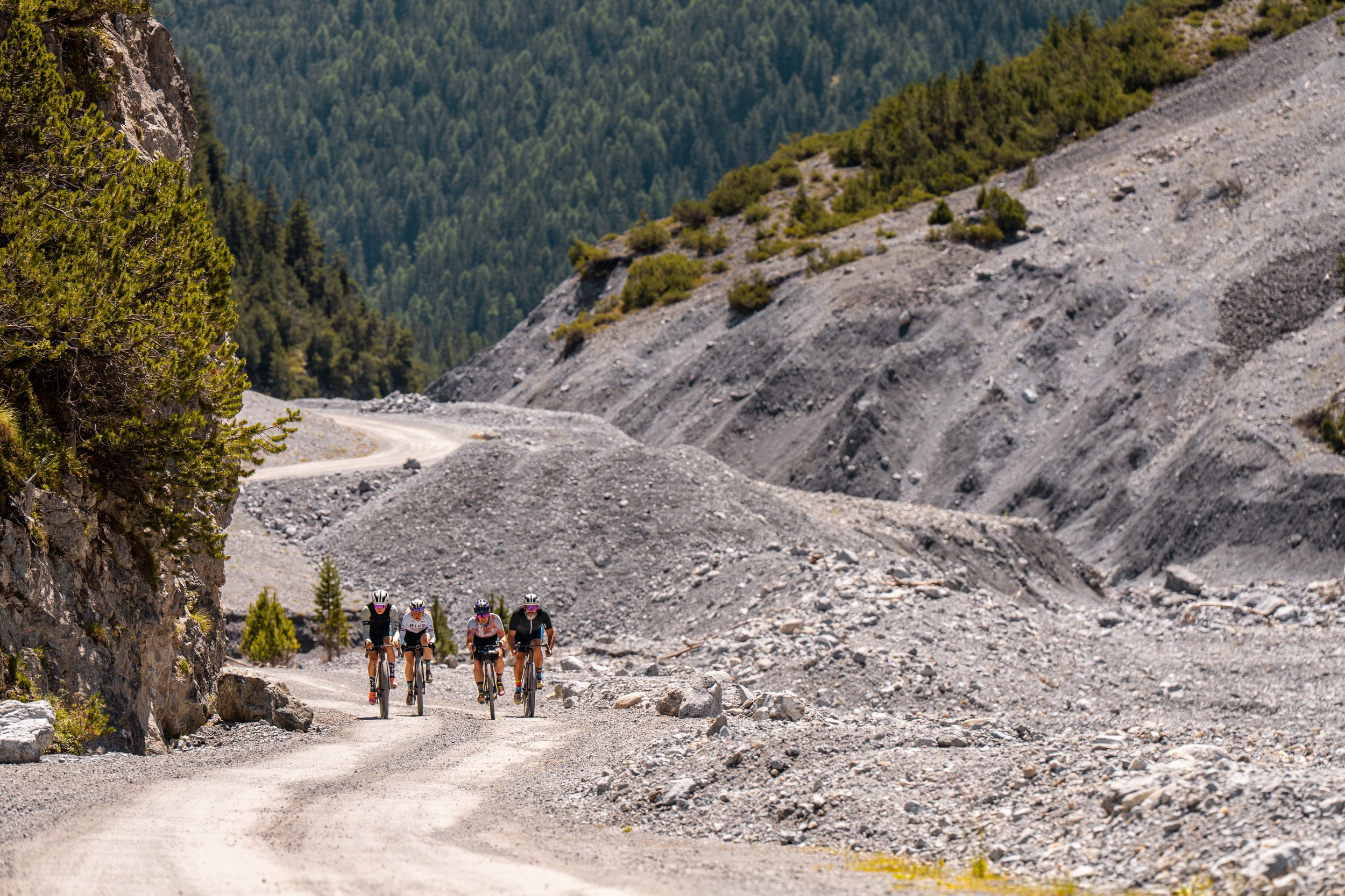 Group of five mountain bikers riding on a dirt trail through rocky and forested mountain landscape with trees and hills in the background.