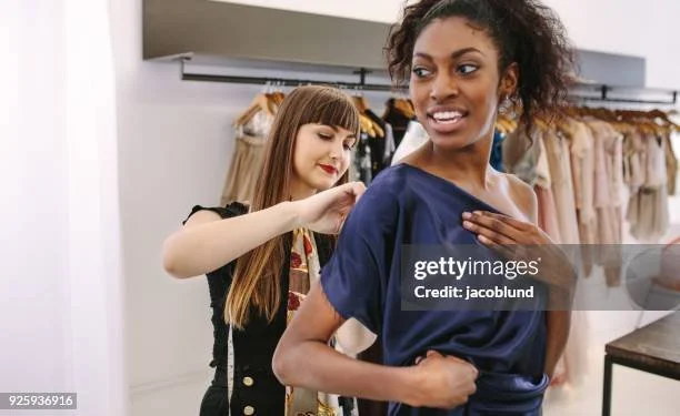 A woman trying on a silky blue top with a woman assisting her in a clothing store.