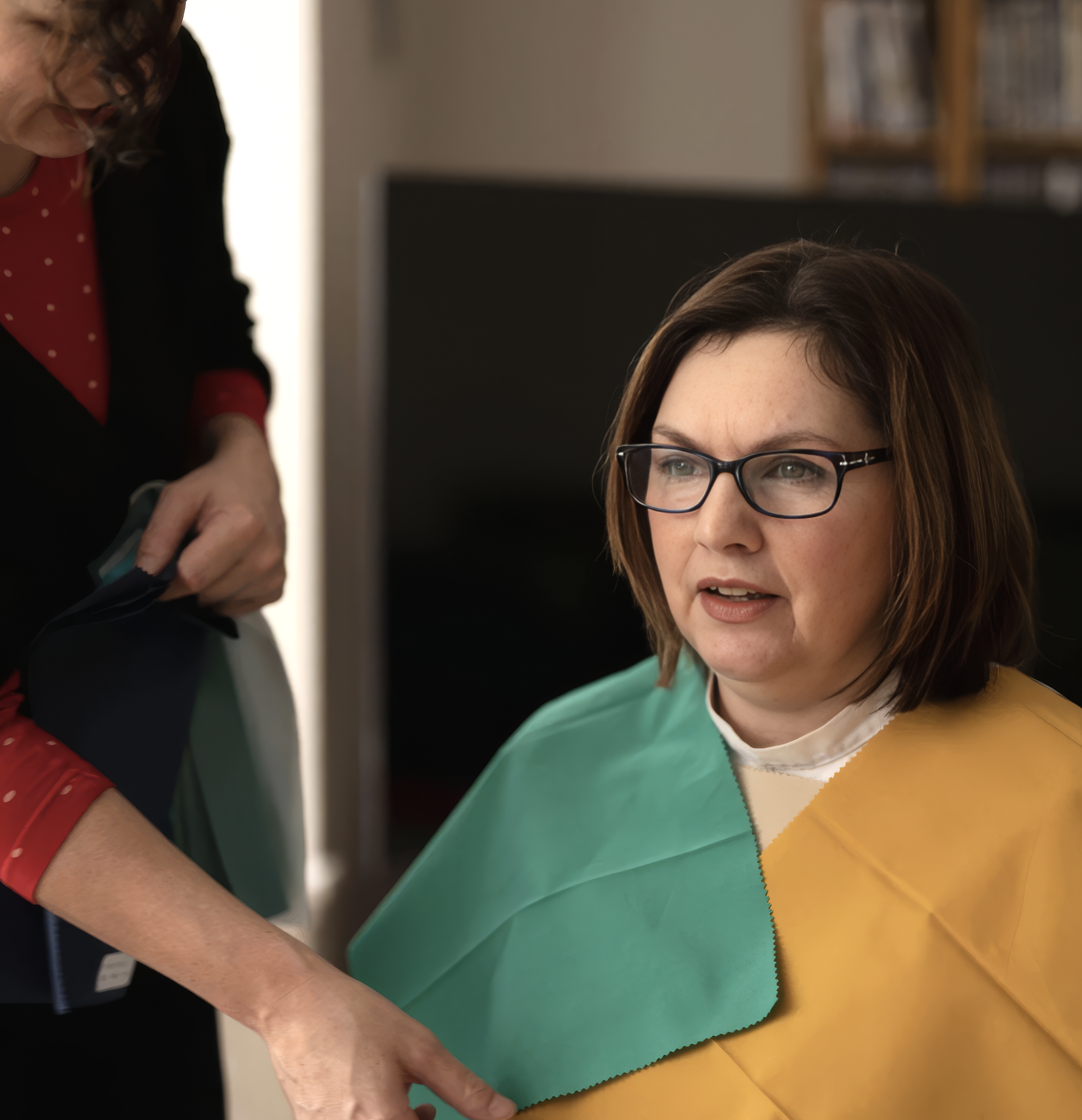 Woman with glasses sitting at a desk with colorful paper in front of her, woman is talking to another person.