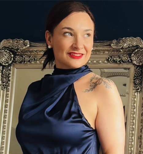 Image of Melanie Foster Lozinski image consultant , in red lipstick, and a tattoo on her left shoulder, wearing a blue satin dress and standing in front of an ornate mirror.