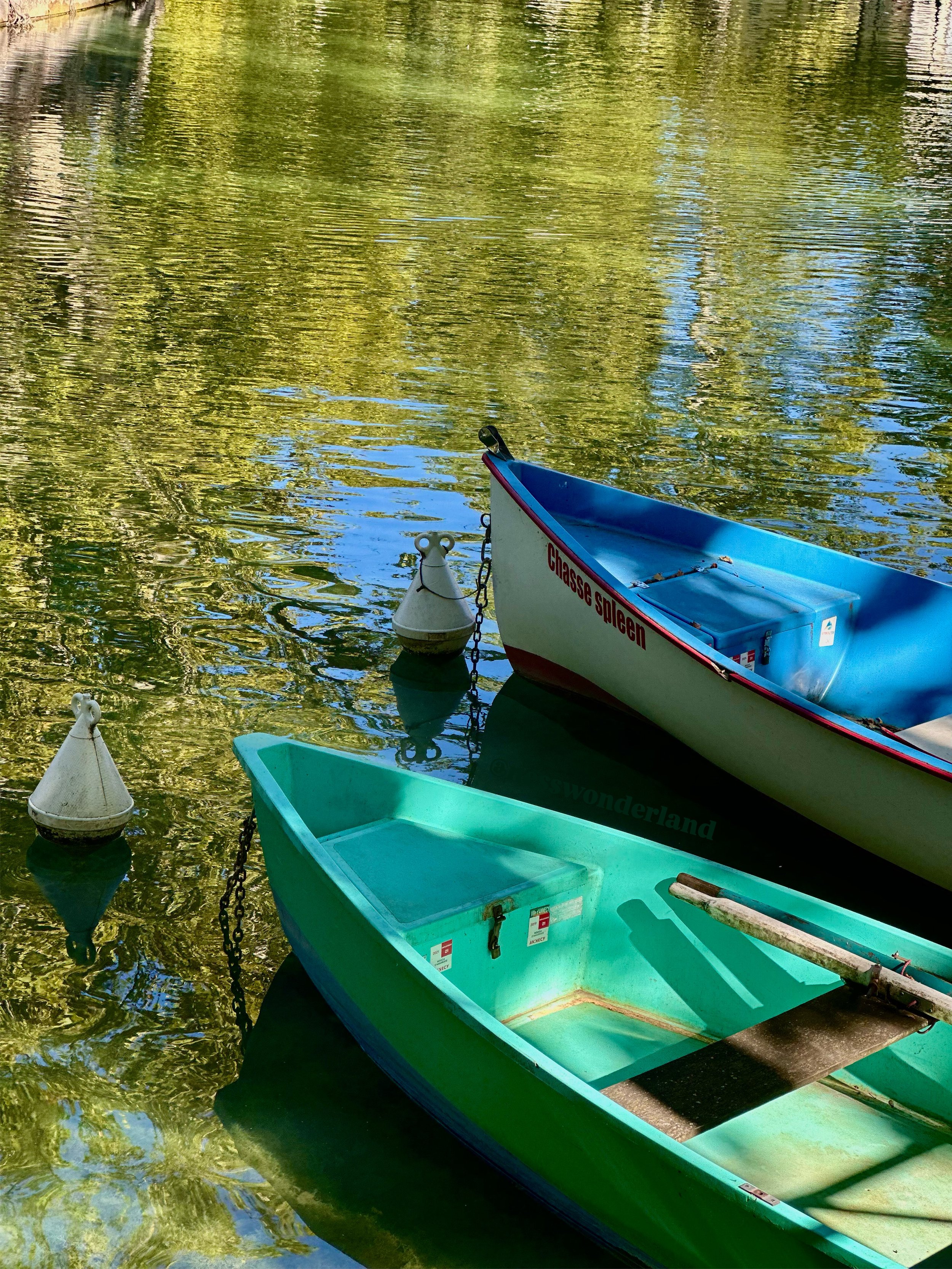 Deux bateaux en plastique, un turquoise et un blanc avec une ligne rouge, amarrés à des bouées dans un lac avec des reflets verts et bleus.