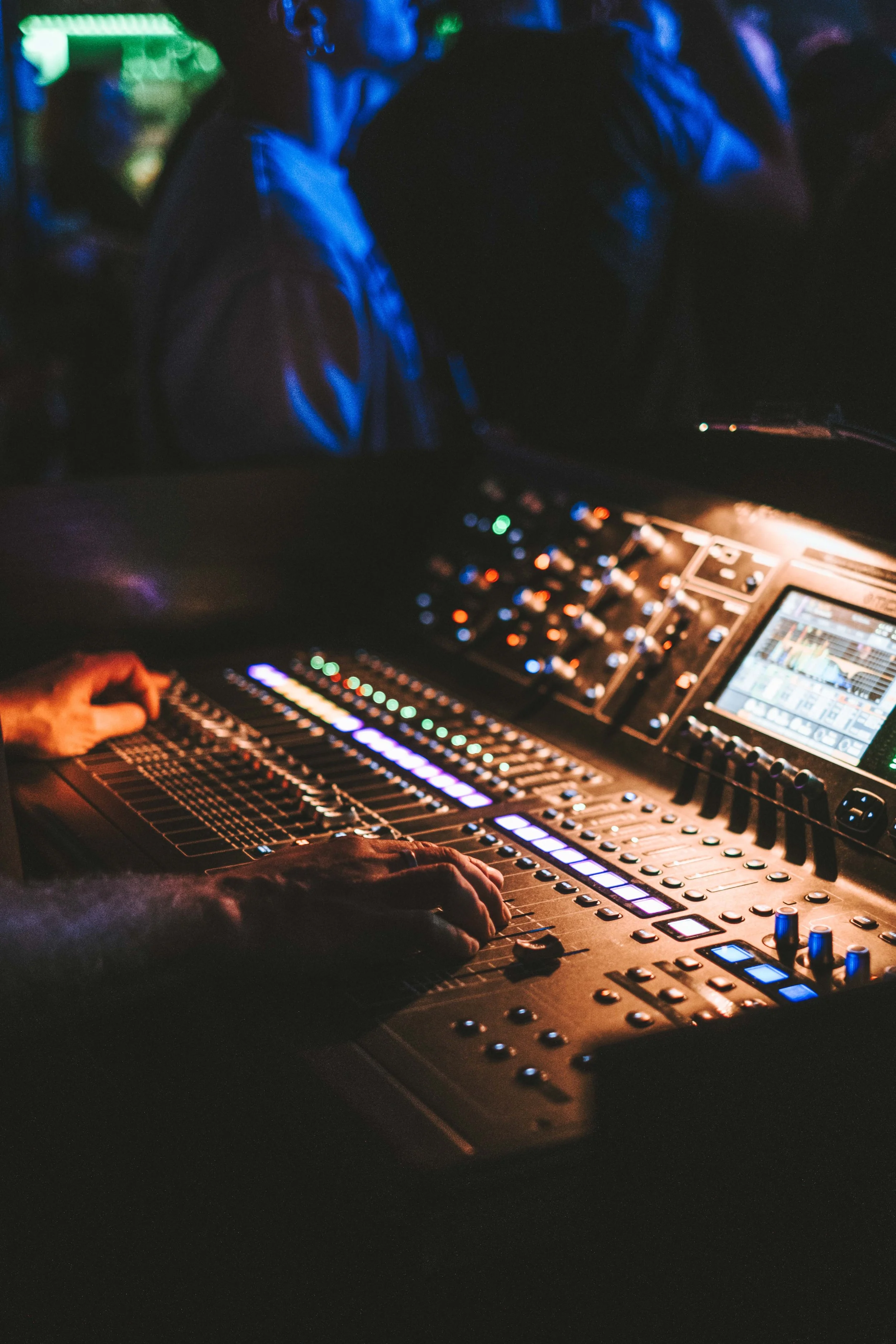 Mischpult mit Händen eines Tontechnikers in einer dunklen Umgebung, möglicherweise in einem Club oder bei einer Veranstaltung, mit leuchtenden Knöpfen und Anzeigen.