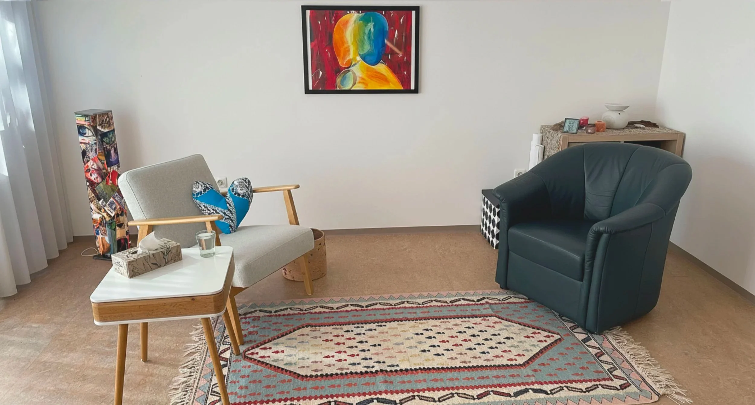 Ein Wohnzimmer mit einem grauen Sessel, einem grünen Ledersessel, einem weißen Beistelltisch, einem bunten Teppich, Wanddekorationen und einer farbenfrohen abstrakten Kunst vor weißer Wand.