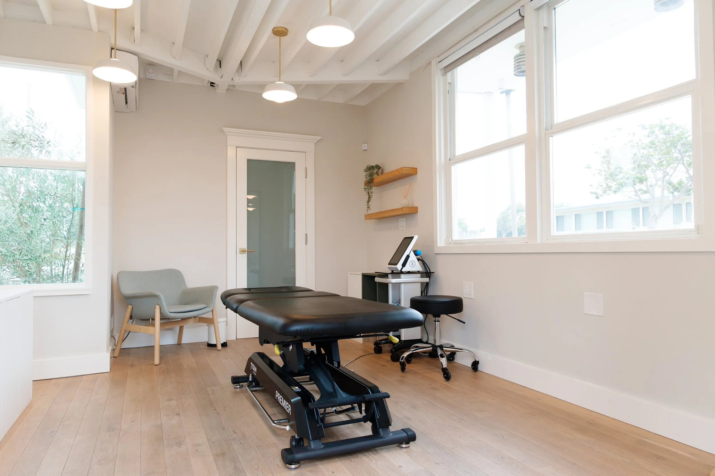 A physical therapy or chiropractic treatment room with a massage table, a chair, and a computer station, featuring large windows and overhead lighting.