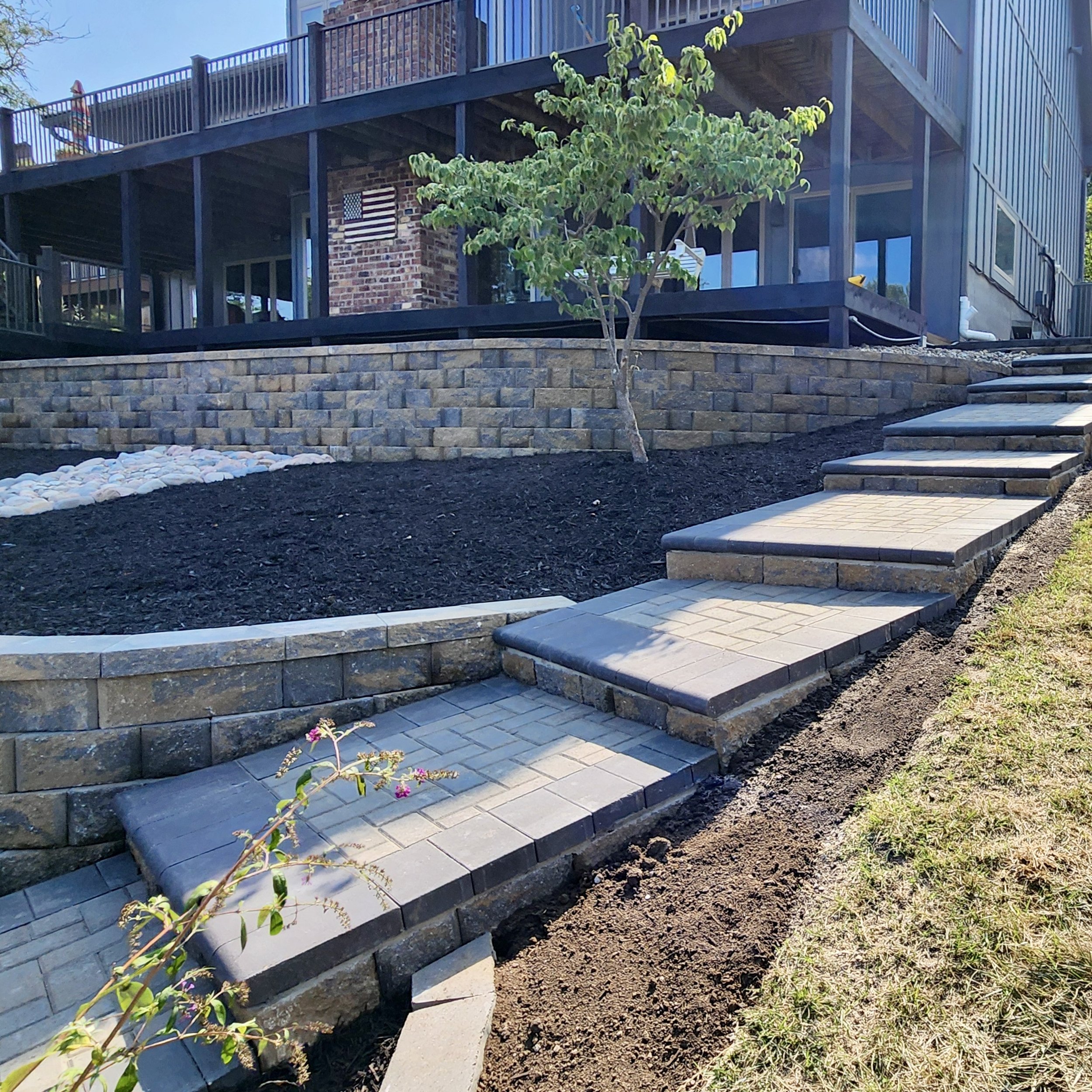 A landscaped backyard with a stone patio and a winding pathway with stairs, a small tree, and a raised brick and stone retaining wall with mulch and decorative white stones.