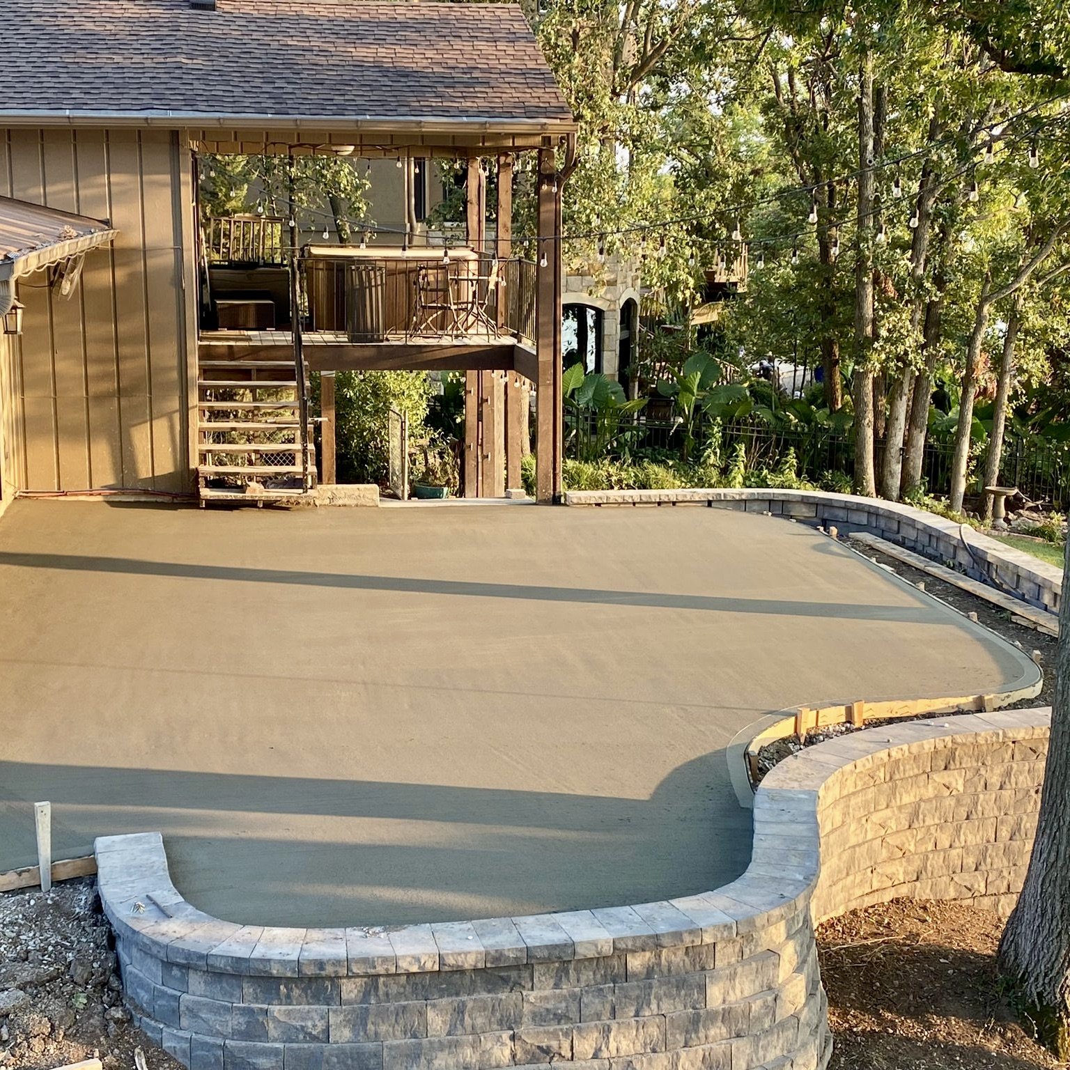 Backyard with a newly paved swimming pool area, a wooden house with a balcony, trees, and string lights.