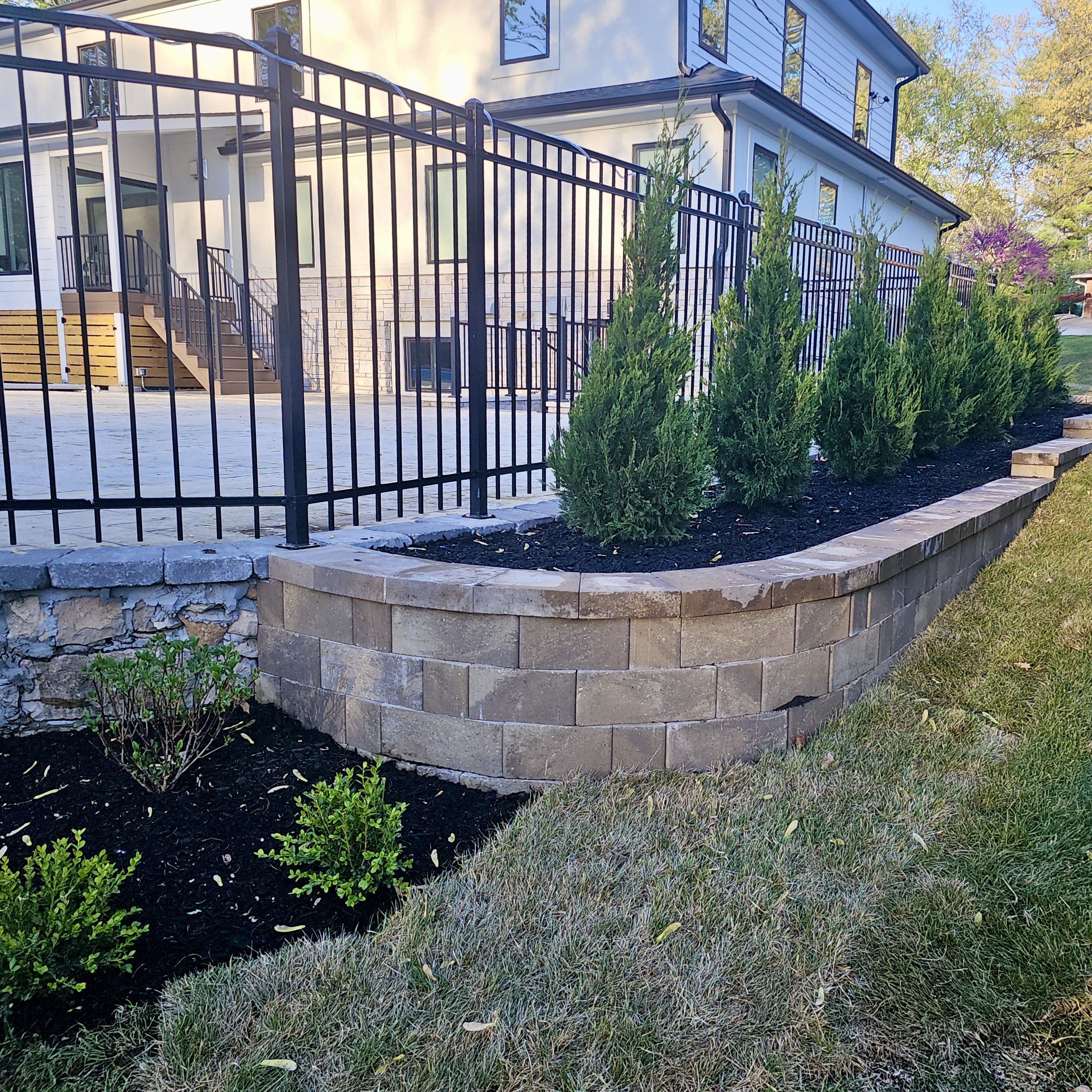 Newly landscaped yard with a brick retaining wall, green shrubs, a black metal fence, and a modern white and blue house in the background.