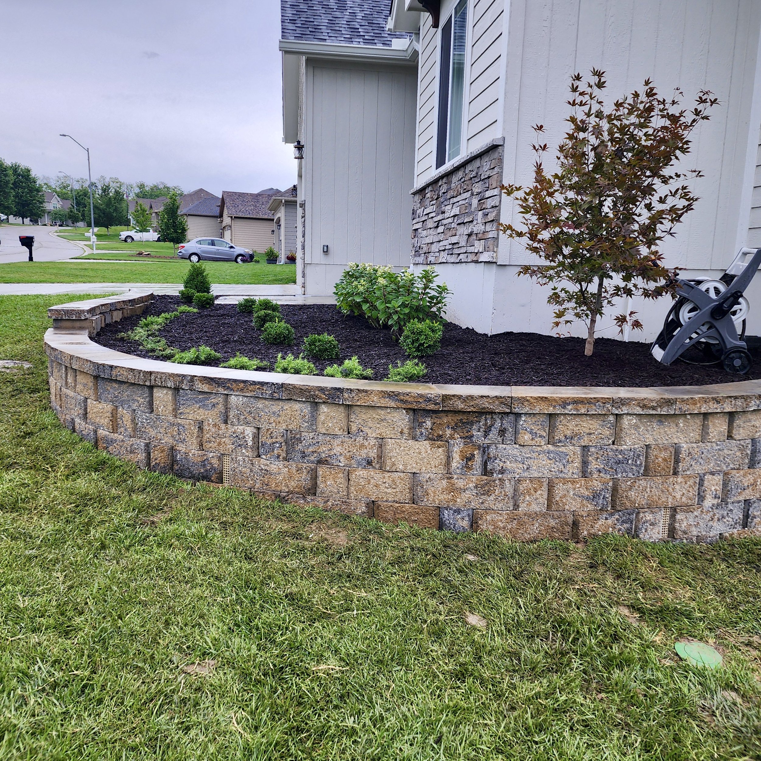 A landscaped front yard with a curved stone retaining wall, green shrubs, a small tree, and a garden hose reel near the house, with a suburban neighborhood in the background.