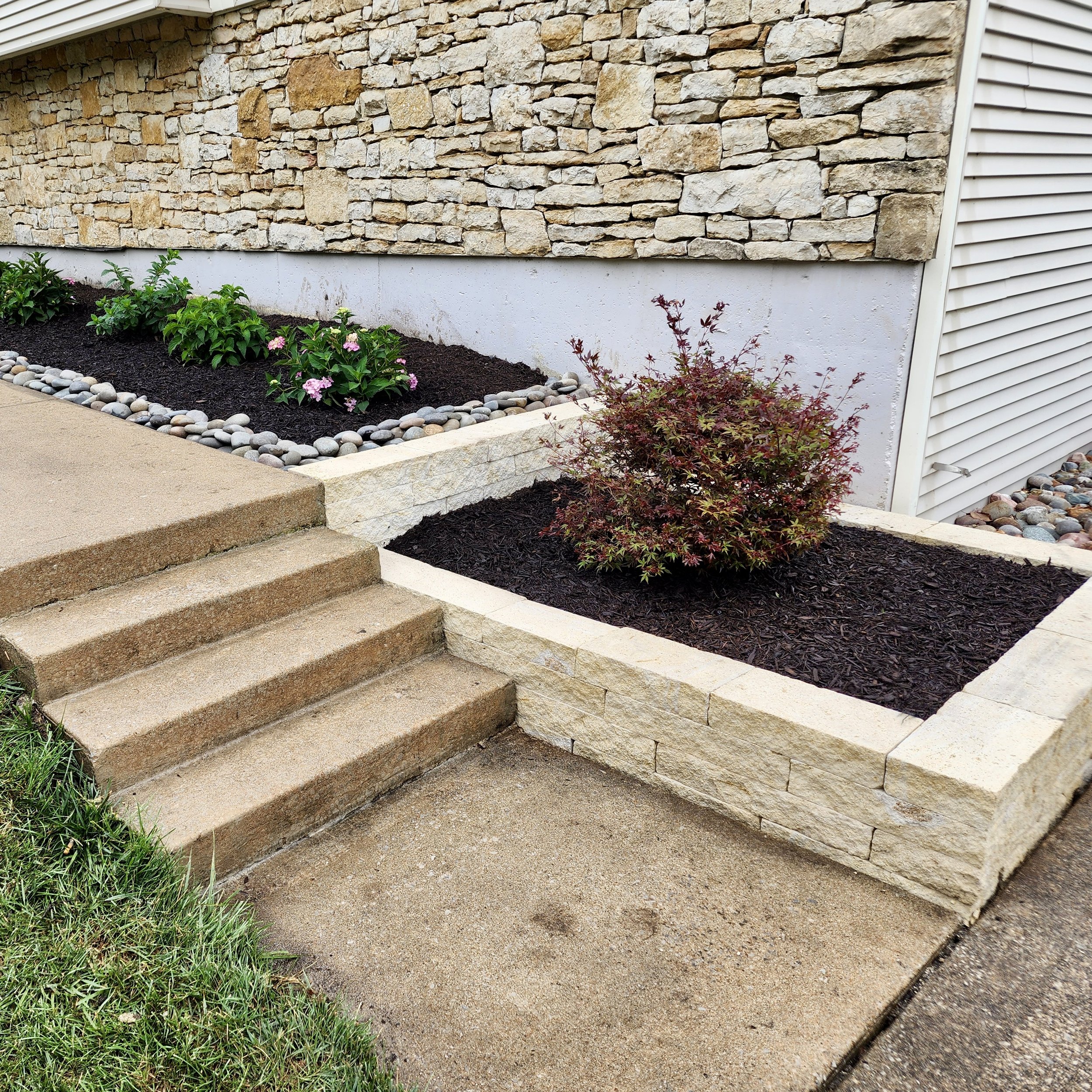 Front yard garden with stone-bordered beds, small shrubs, flowers, and steps leading to the sidewalk.