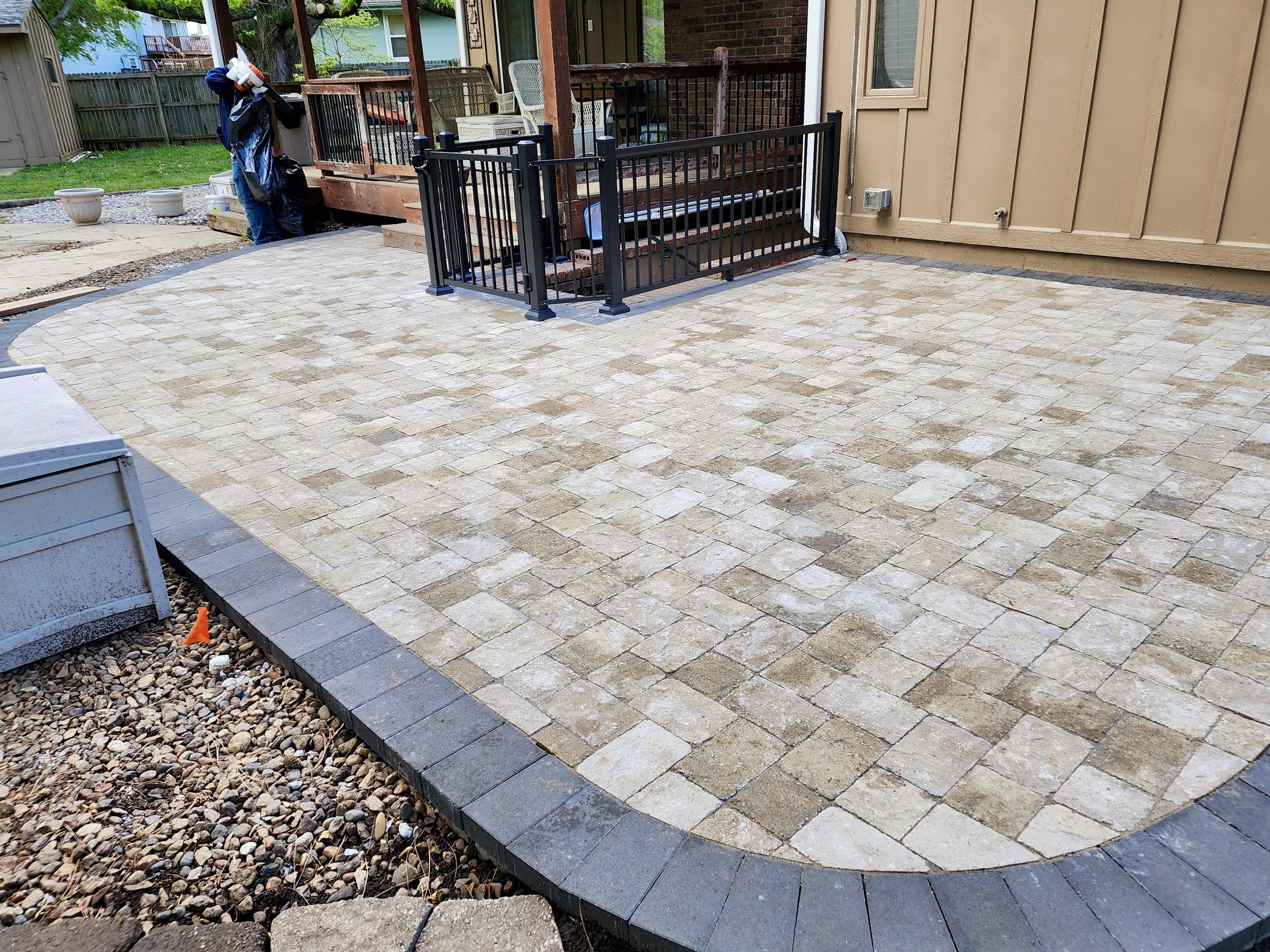 A newly paved backyard patio with beige and tan pavers, a black metal rail at the stairs leading up to a deck, and a person working on the yard in the background.