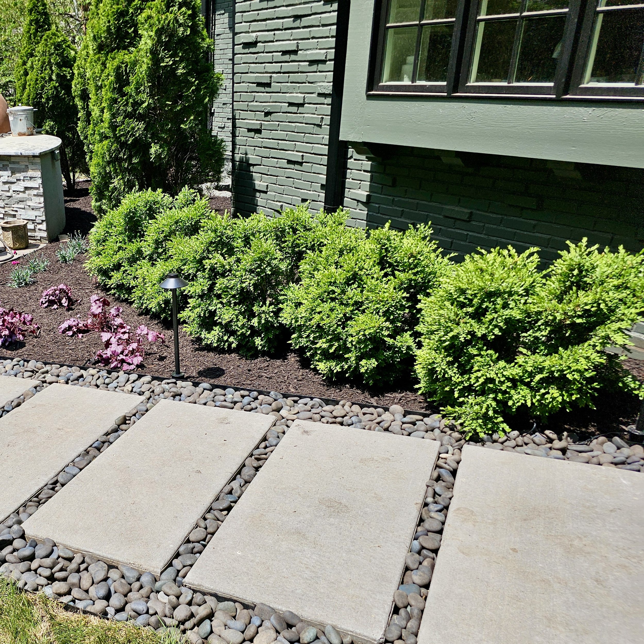 A landscaped area outside a house with green bushes, pink flowers, a small solar light, stone pavers, and mulch near a gray stone and brick house with a window.