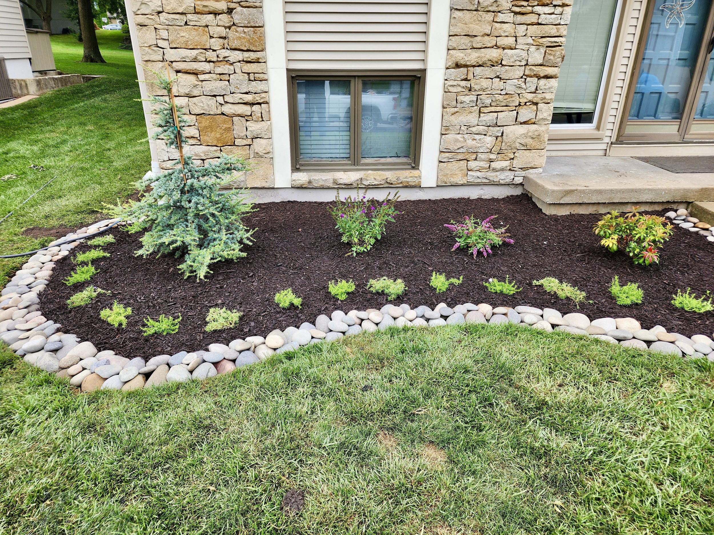 A small garden bed with young plants, mulch, and a stone border in front of a house with brick and siding exterior.