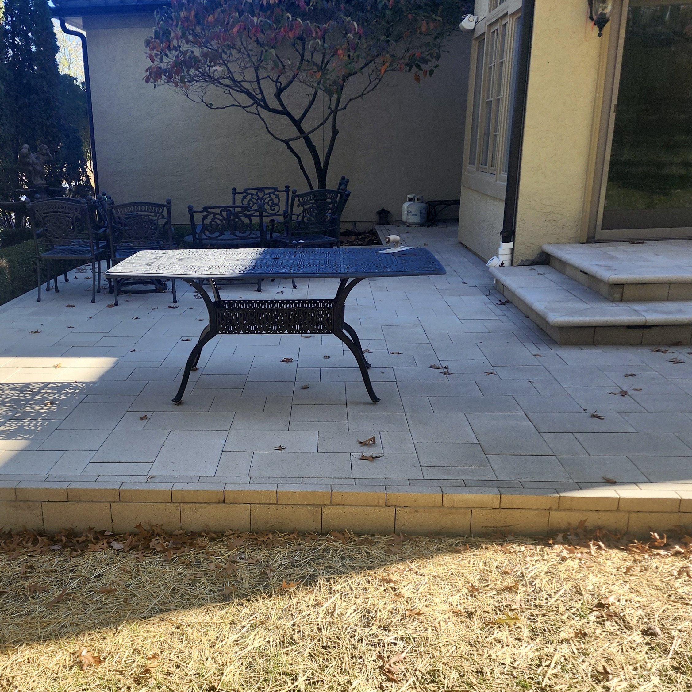 An outdoor patio area with a rectangular table and four chairs, a tree with red leaves, and steps leading to a house with a large window. The patio is paved with light-colored tiles, and there are some fallen leaves scattered across it.