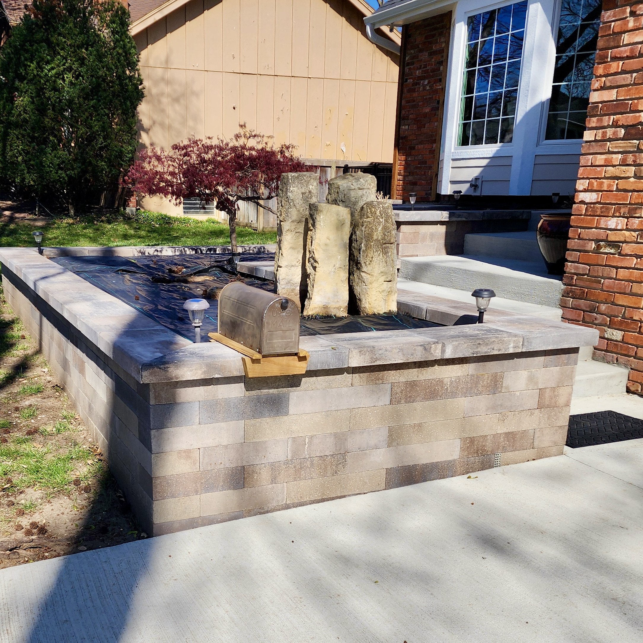 Front yard with brick and stone planter bed, small tree, mailbox, and steps leading to house entrance.