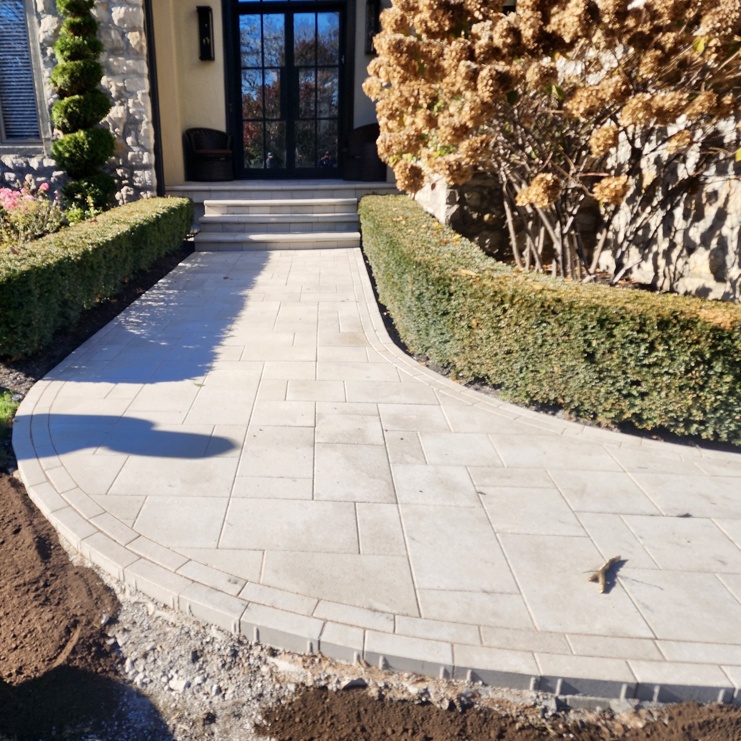 Paved stone walkway leading to front steps of house, flanked by neatly trimmed bushes and a flowering tree on the right, with the house entrance visible in the background.