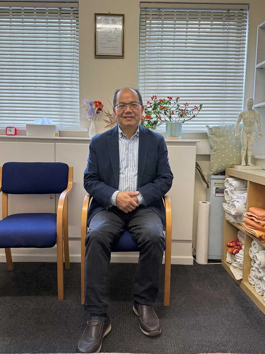 A man in glasses, a blue blazer, a collared shirt, and dark pants sitting on a wooden chair in an office or clinic setting. Behind him are two windows with blinds, a shelf with folded towels, a model of the human body, and decorative plants.