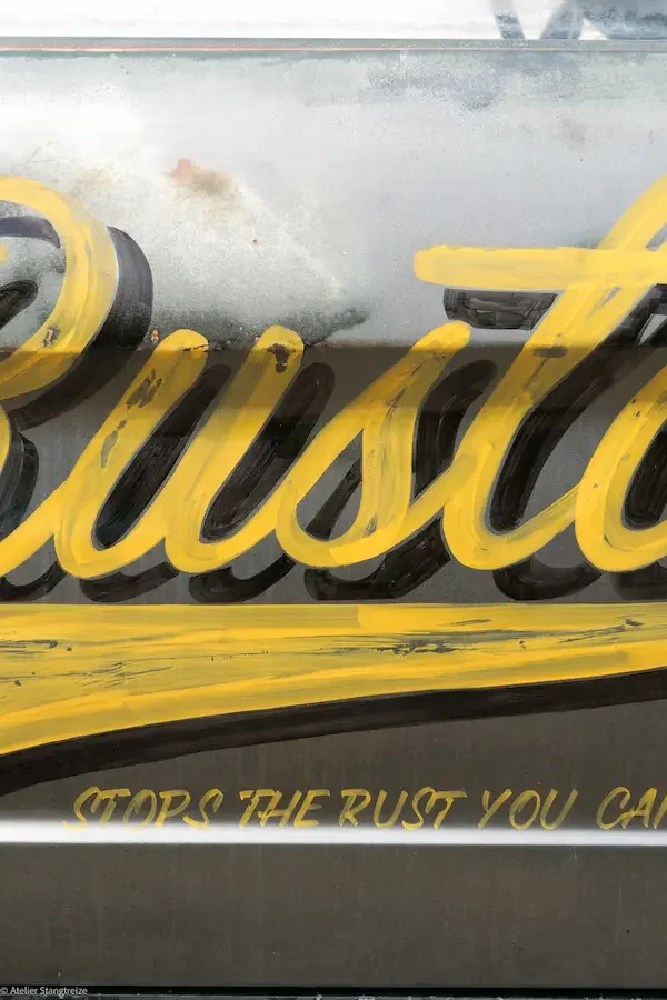 le mot "Rustol"  en lettres jaunes cursives peint sur une carrosserie usée de Ford Mustang, accompagnée du slogan « STOPS RUST YOU CAN'T SEE ».
