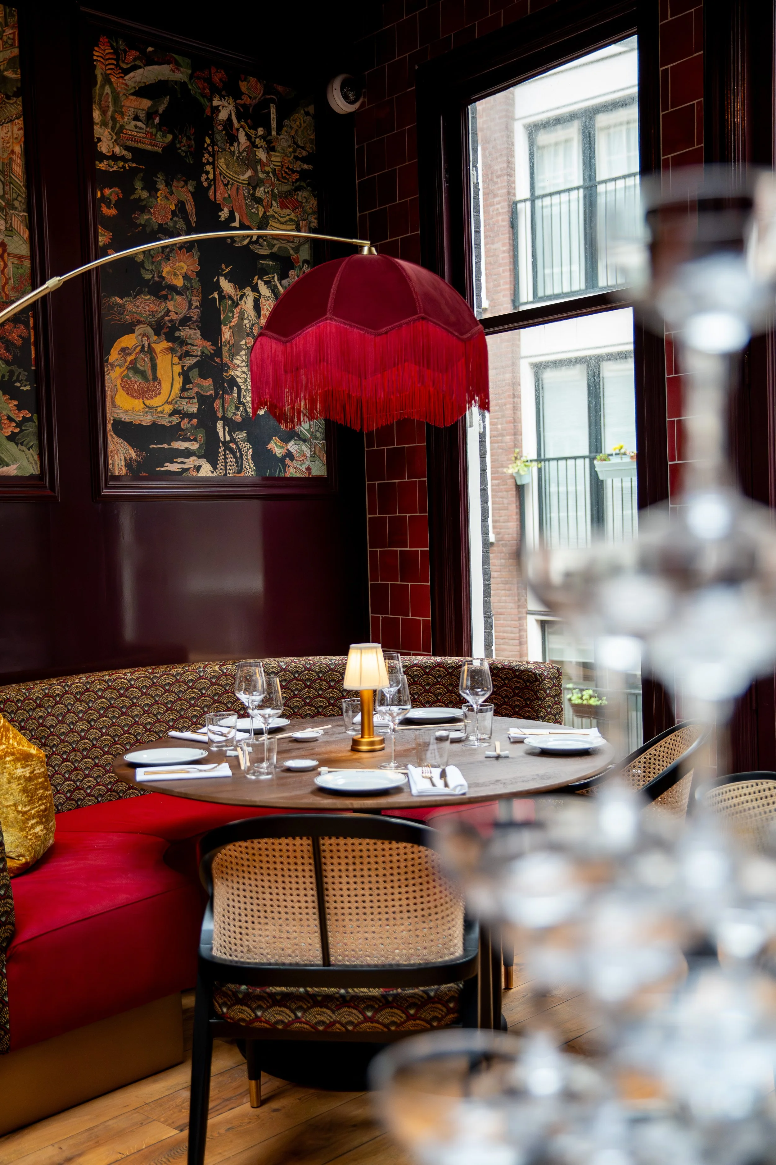 Een elegante eetzaal met een ronde tafel, gedekt voor diners, omgeven door gemengde stoelen. Een grote rode lampenkap hangt boven de tafel. Een geluidsisolerende muur met een traditioneel patroon en ramen die uitkijken op een buitenbalcony. Een tafellamp in het midden van de tafel.